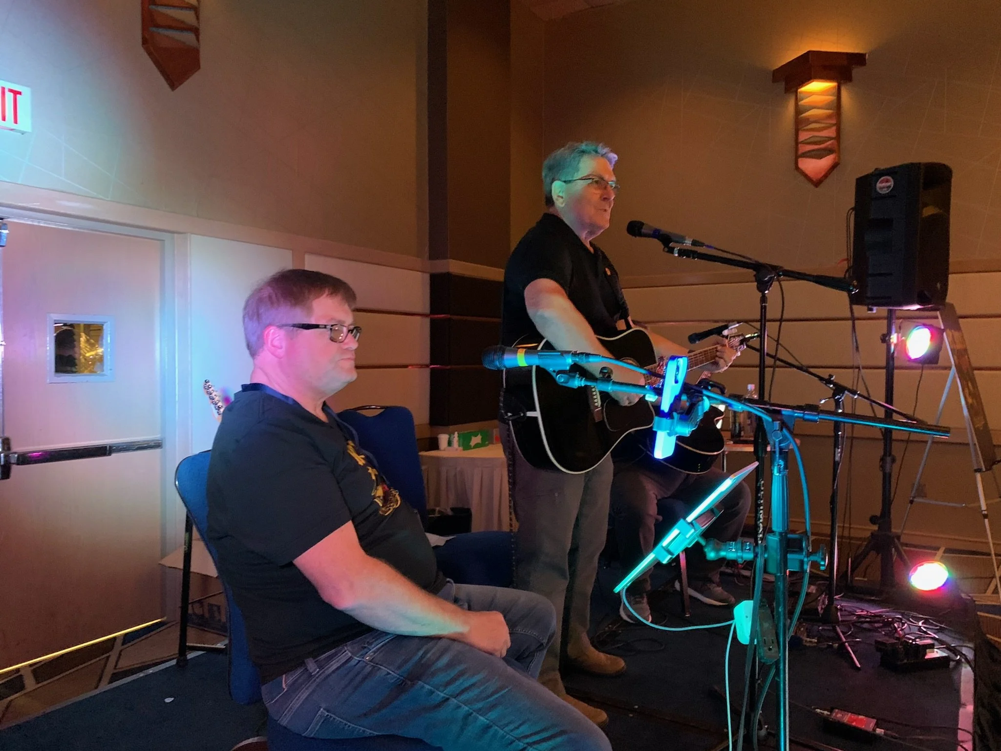 Two men performing music on stage, one playing guitar and standing, the other sitting with glasses, in a dimly lit indoor venue.