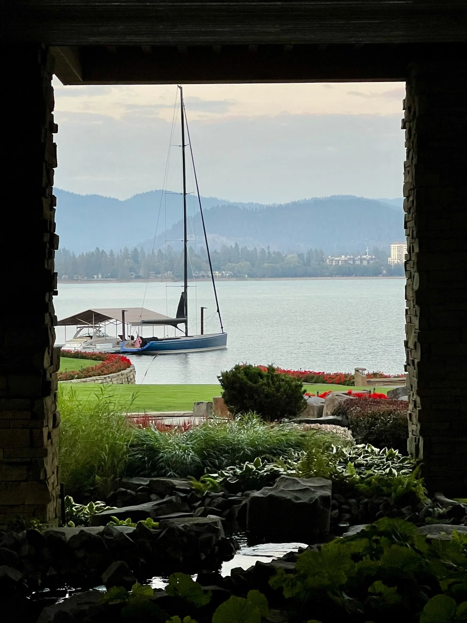 View of a lake with a sailboat and a motorboat, taken from under a stone archway with a garden in the foreground and mountains in the background