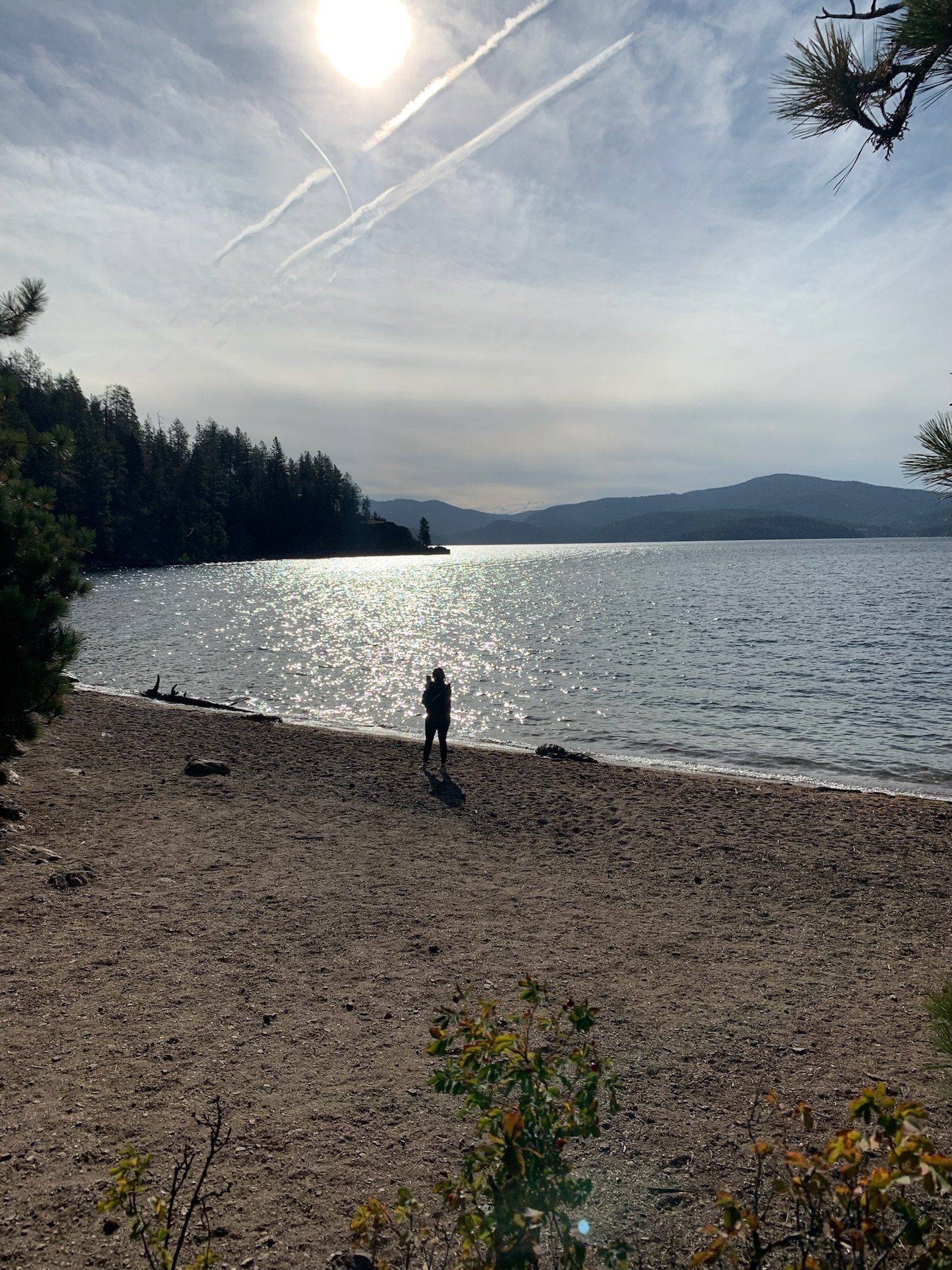 Person standing on a beach, taking a photo, with the sun reflecting off the water during daytime. Trees frame the scene, mountains in the background, and the sky has contrails.