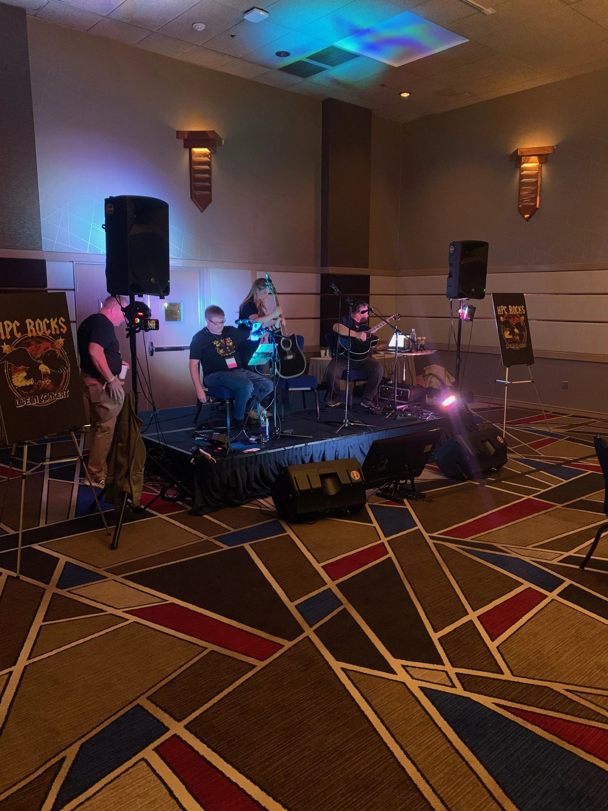 A small musical band with three members performing on a stage in a hotel conference room, with colorful lighting.