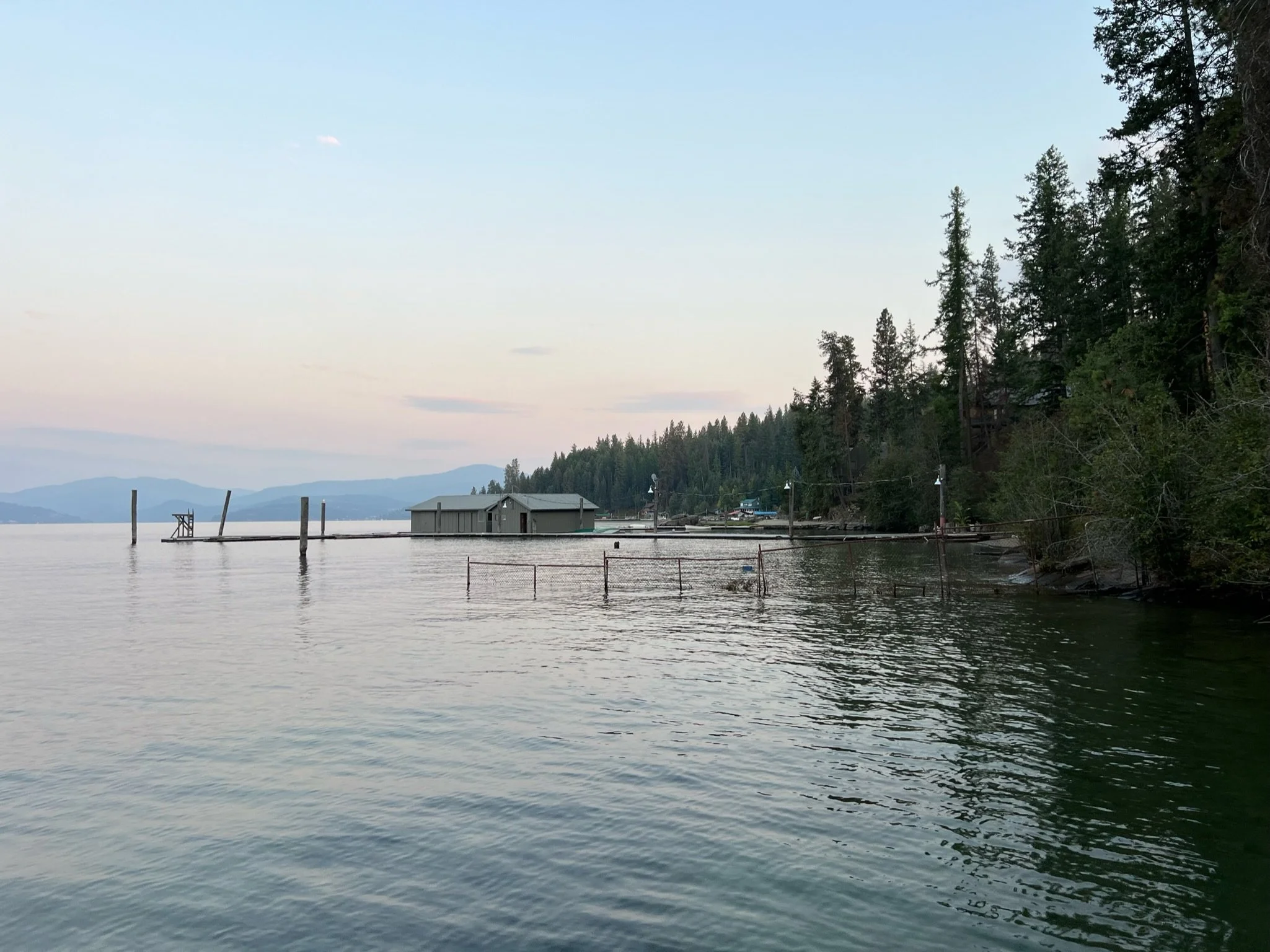 A peaceful lakeside scene at sunset with calm water, a small building near the shore, wooden docks extending into the lake, and dense evergreen trees on the right side.
