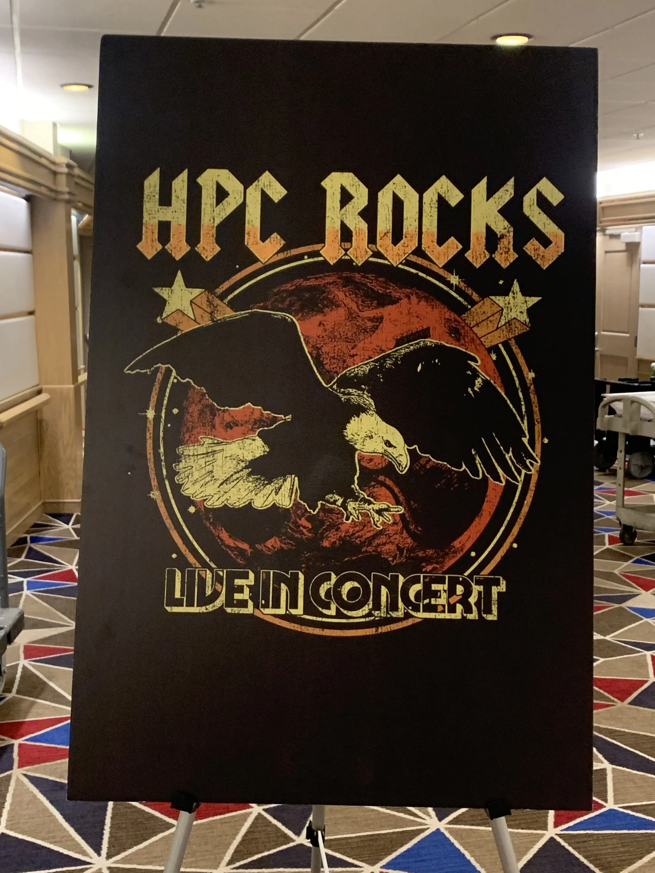 Concert sign with a black background. Features an illustration of a bald eagle with wings spread, set against an orange moon. Text at the top reads 'HPC ROCKS'. Text at the bottom reads 'LIVE IN CONCERT'.