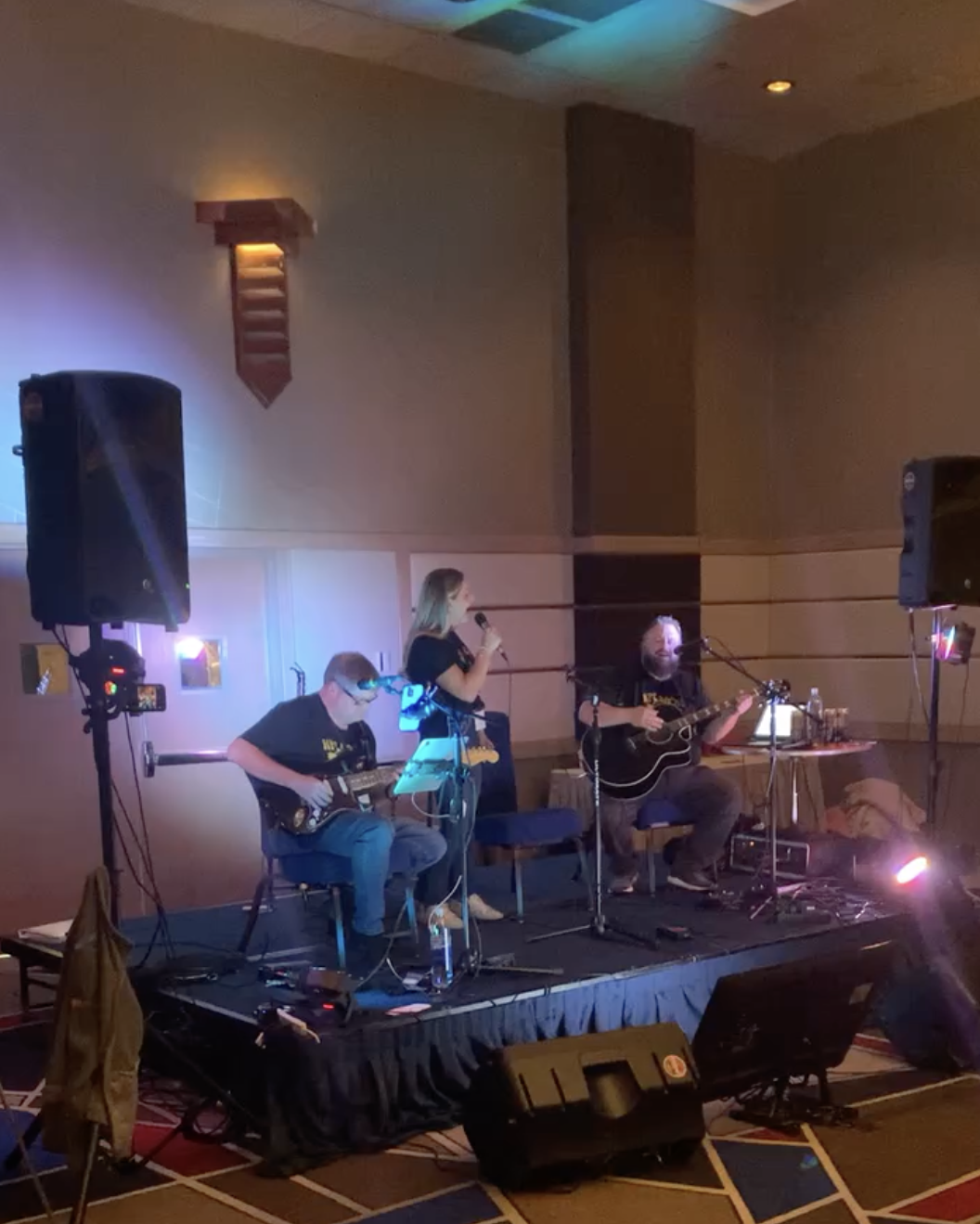 A band performing live on a stage in a dimly lit room, with musicians playing guitars and a woman singing into a microphone.