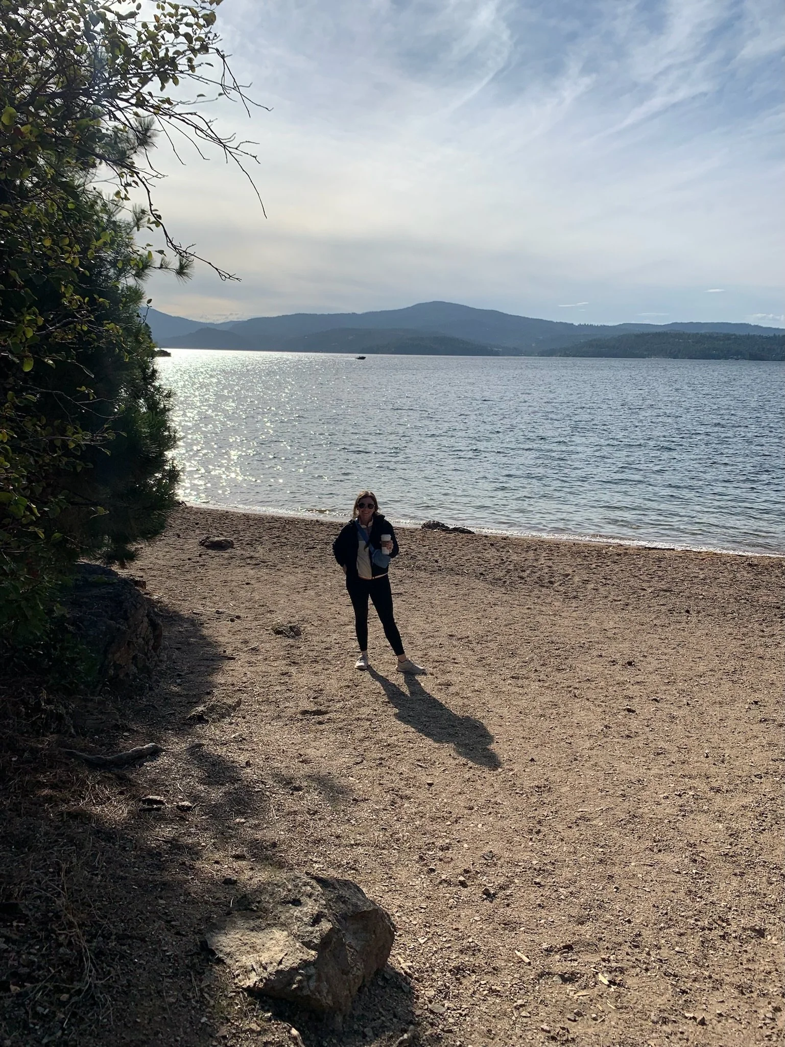 A woman standing on a sandy beach by a body of water with mountains in the distance, sunlight reflecting off the water, and a partly cloudy sky.