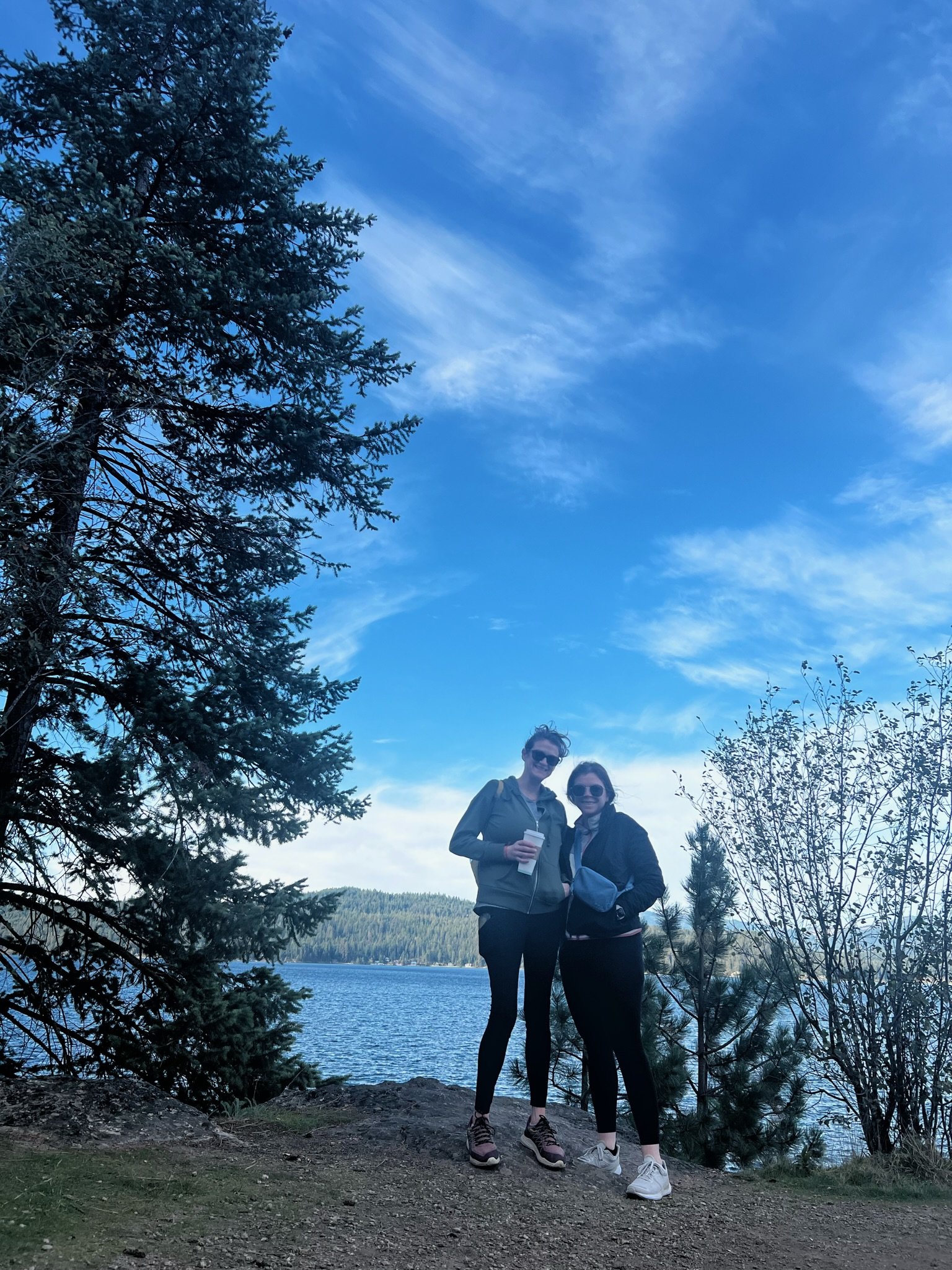 Two women standing by a lake surrounded by trees, with a blue sky and clouds above.
