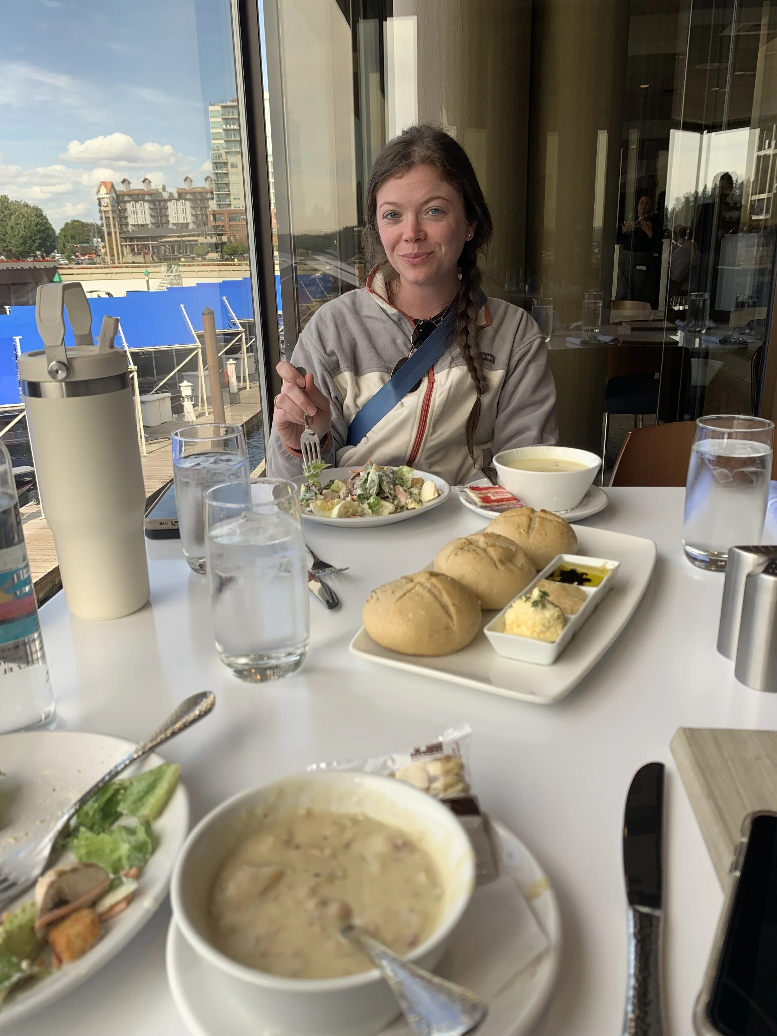 A woman with long brown hair in a braid, sitting at a table in a restaurant near a window with a view of downtown buildings, smiling and holding a fork. The table has salads, bread rolls with butter and olive oil, and bowls of soup, with glasses of w