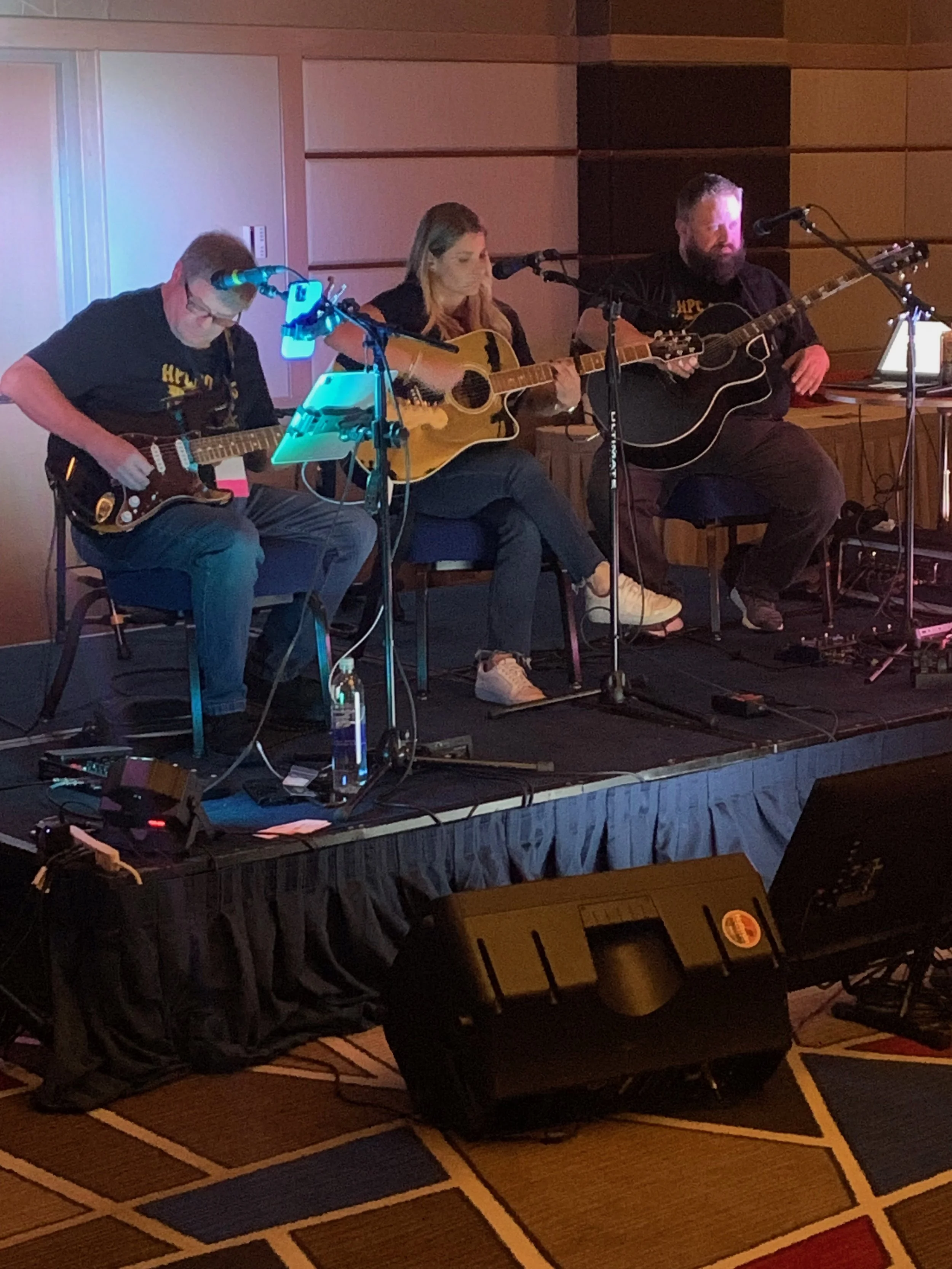 Three musicians playing acoustic guitars on a small stage indoors, with sound equipment and a black curtain in the background.