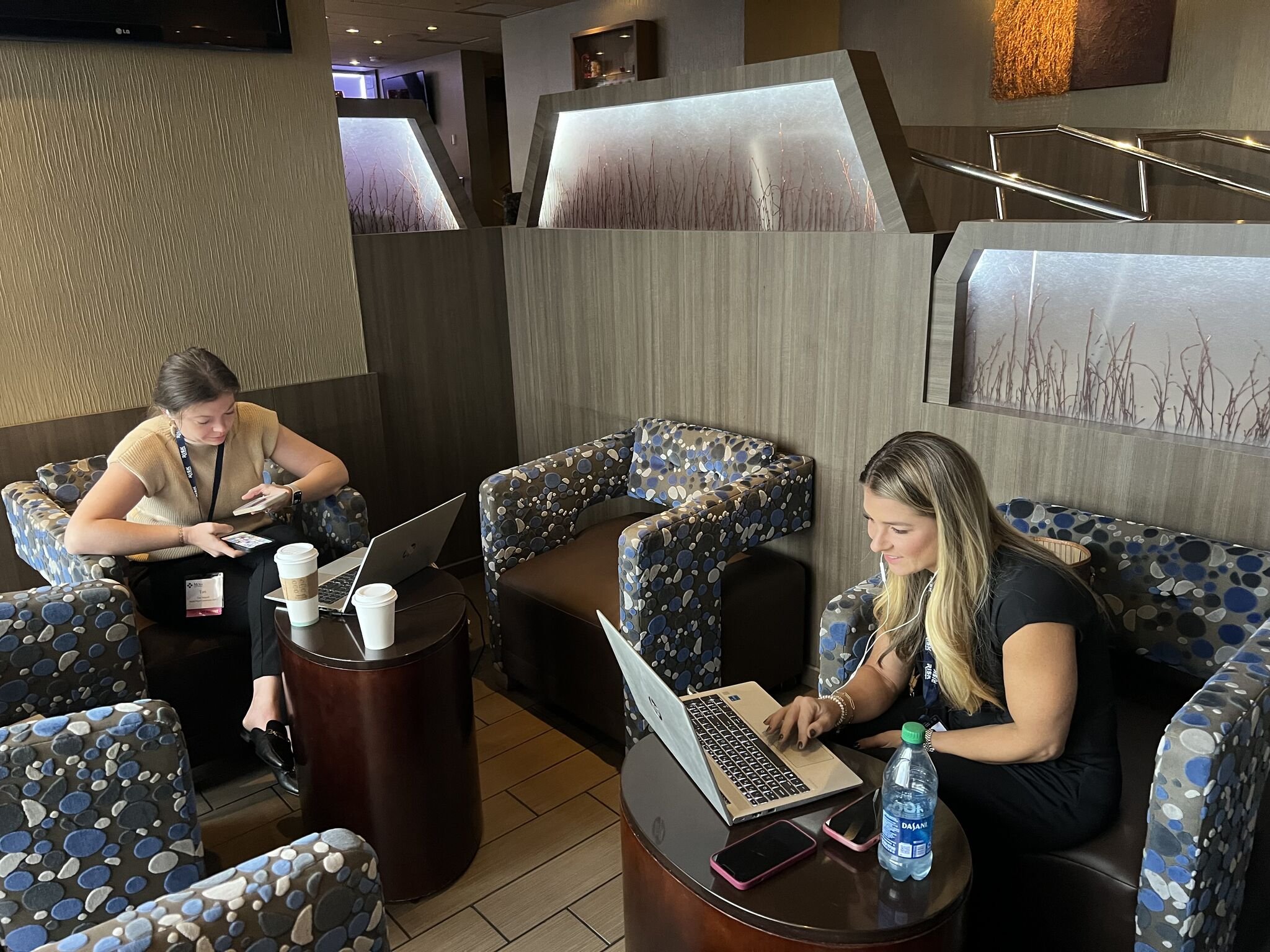 Two women working on laptops in a lounge area with patterned chairs and coffee cups on tables.