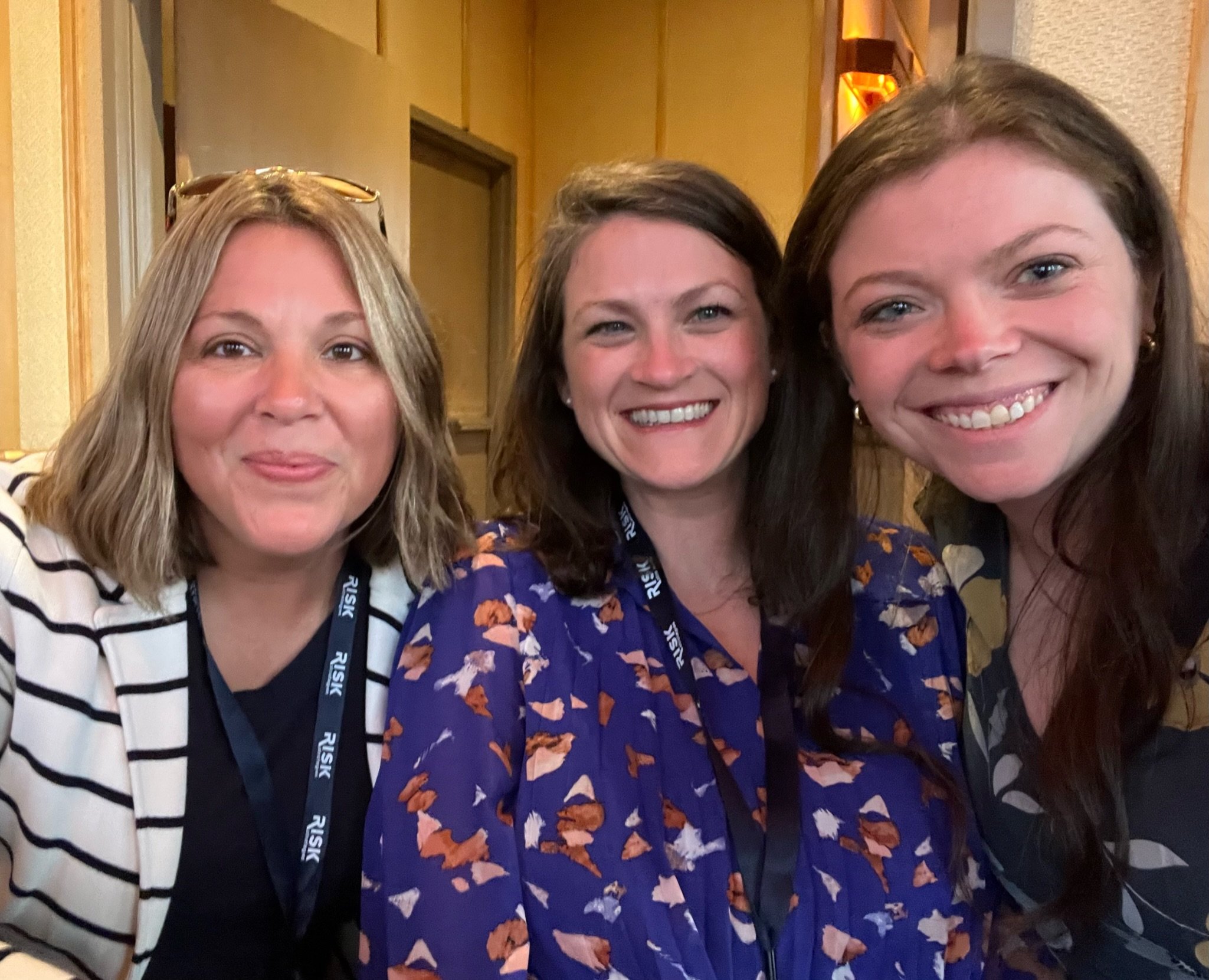 Three women smiling for a selfie indoors with wooden walls and warm lighting in the background.