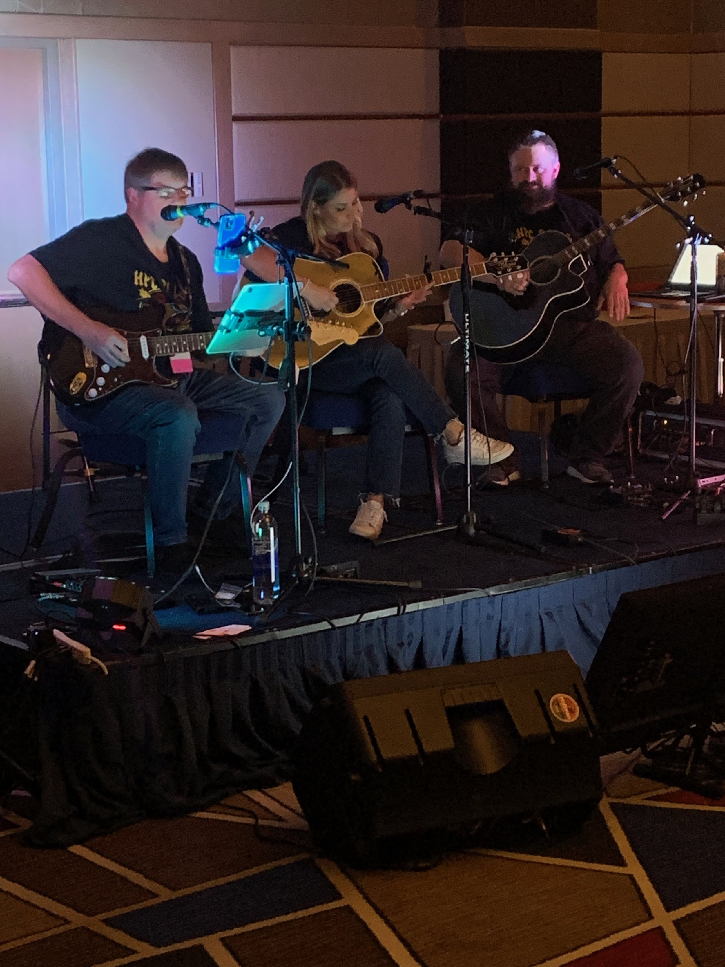 Three musicians performing on stage: a young man with glasses playing an electric guitar, a woman with long hair playing an acoustic guitar, and a man with a beard playing an acoustic guitar, with microphones and audio equipment around them.
