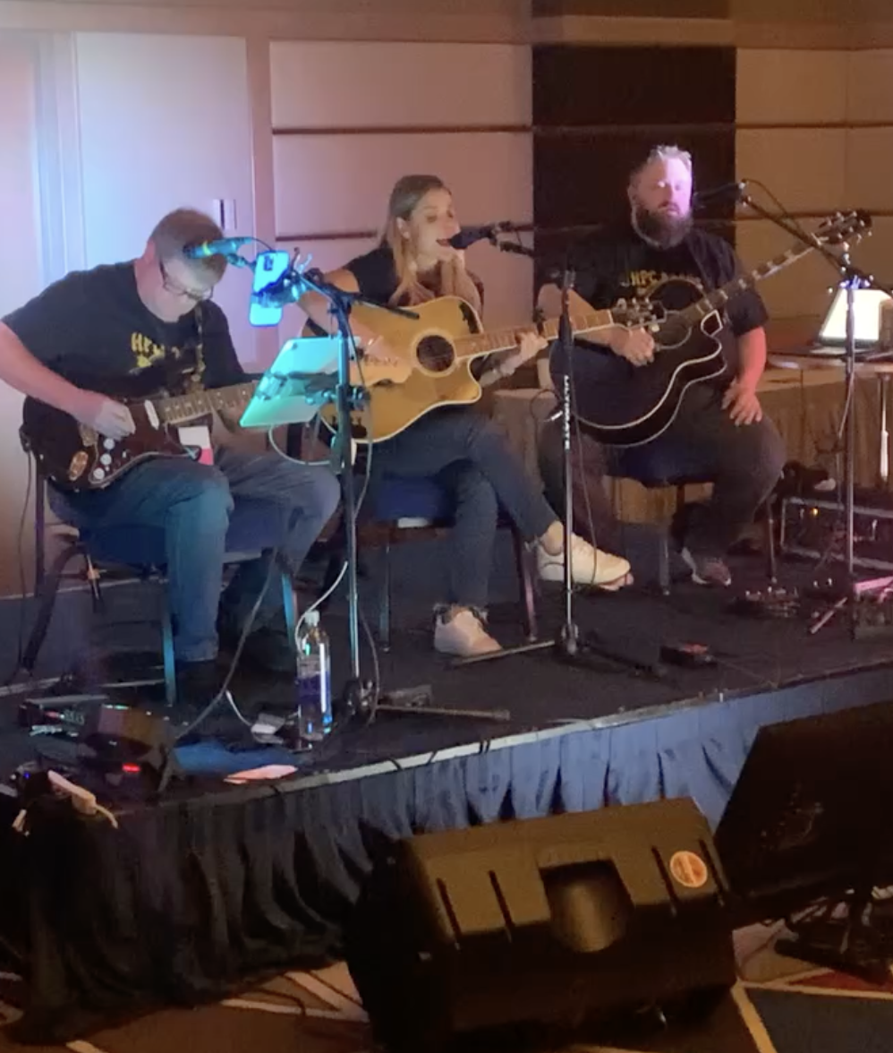 Three musicians performing on a small stage with guitars and microphones, in a cozy indoor setting.
