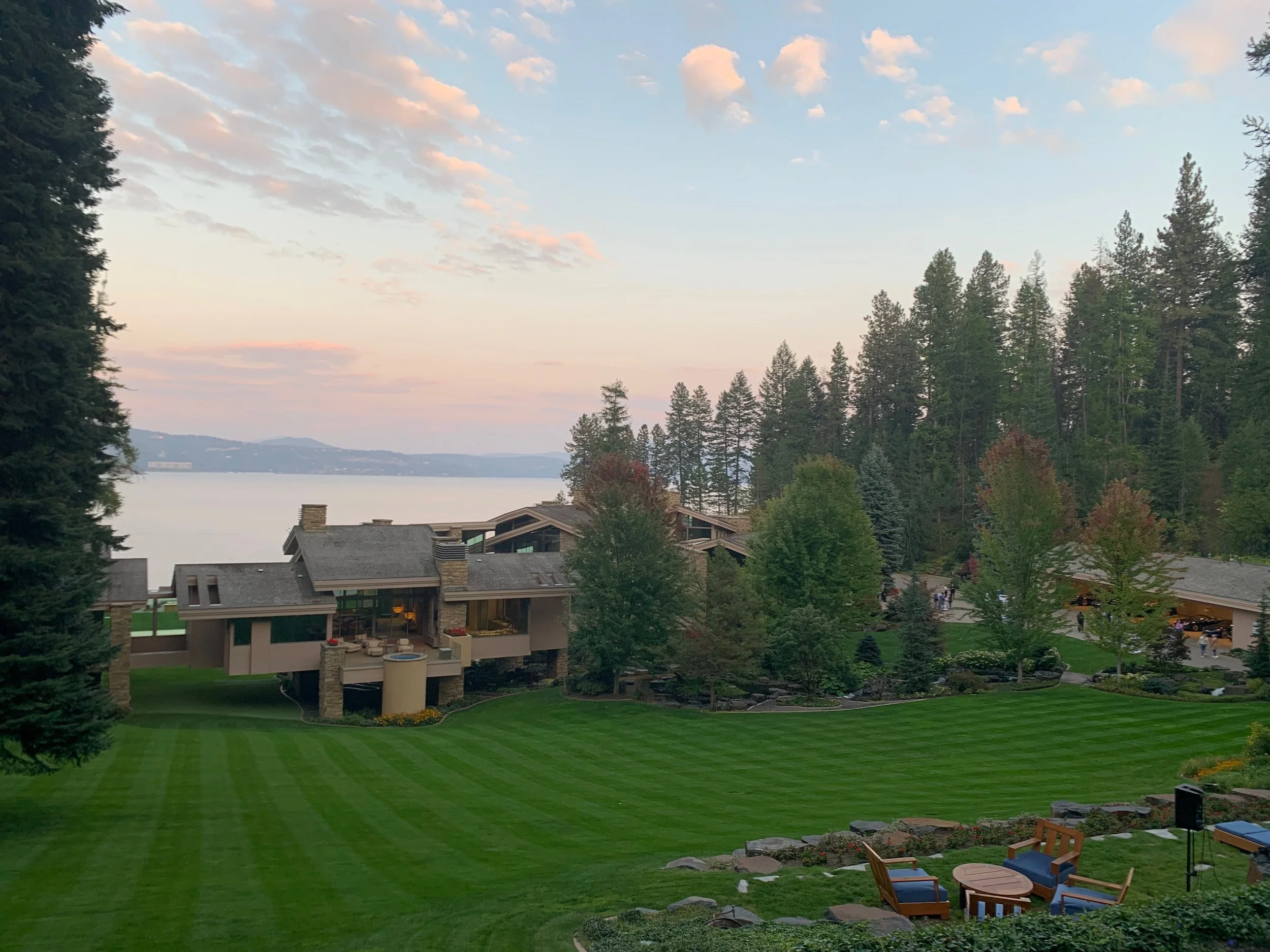 A scenic view of a modern house with a spacious patio overlooking a large lake, surrounded by lush green trees and a well-manicured lawn with outdoor furniture.