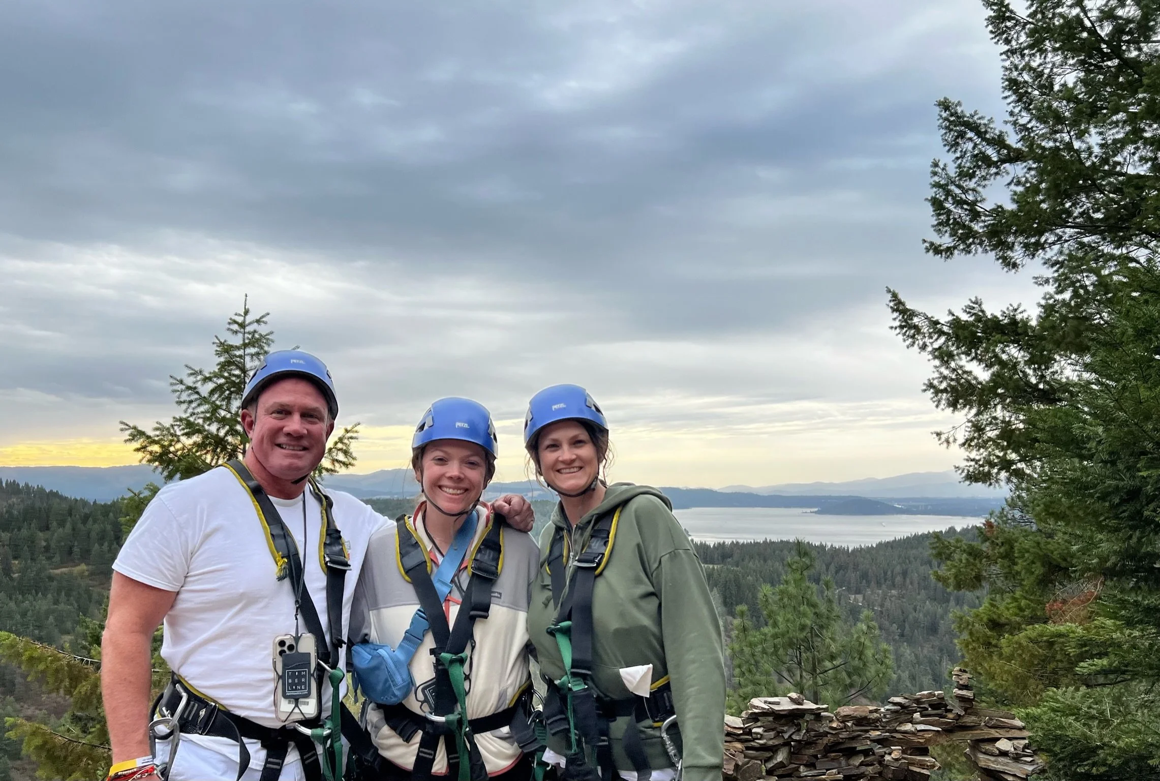 Three people wearing helmets and harnesses, standing on a viewpoint with a scenic forest and body of water in the background.