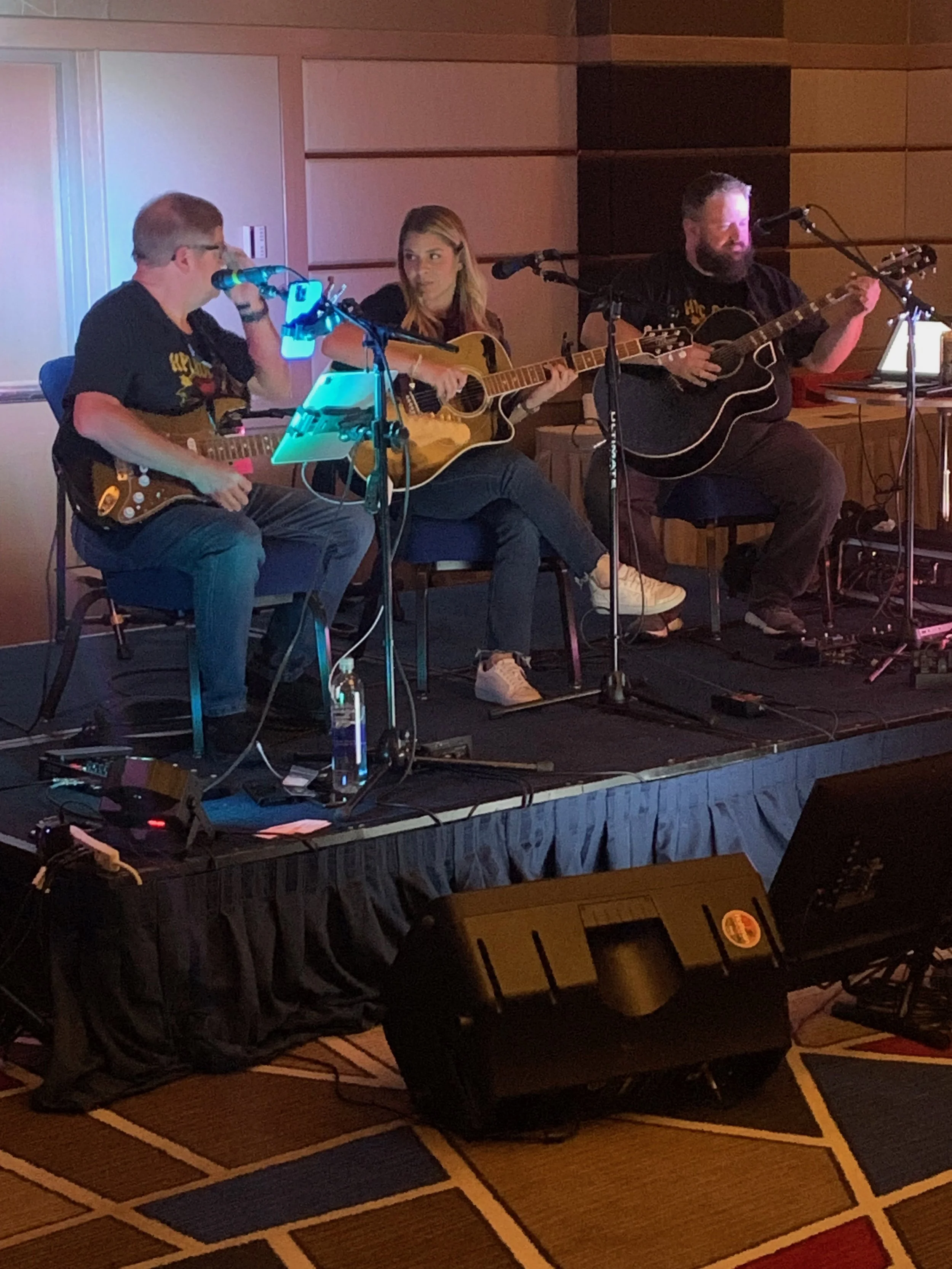 Three musicians playing guitars on a stage, performing indoors with microphones and sound equipment, under warm lighting.