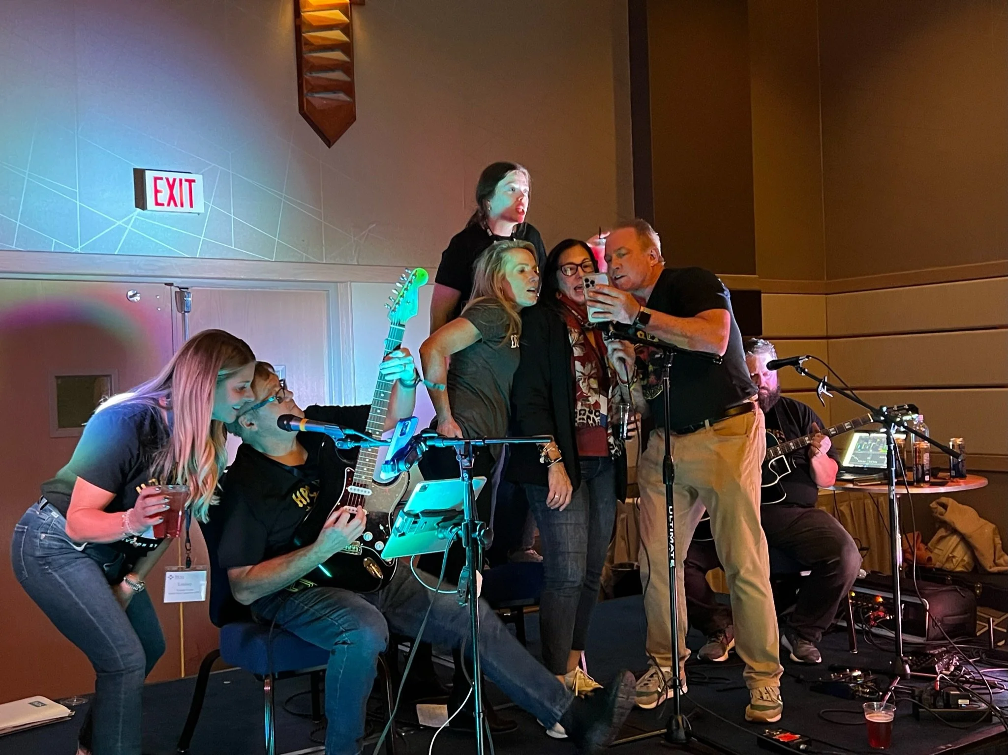 Group of people on stage during karaoke, some holding guitars and microphones, with one person taking a selfie, in a room with an exit sign in the background.