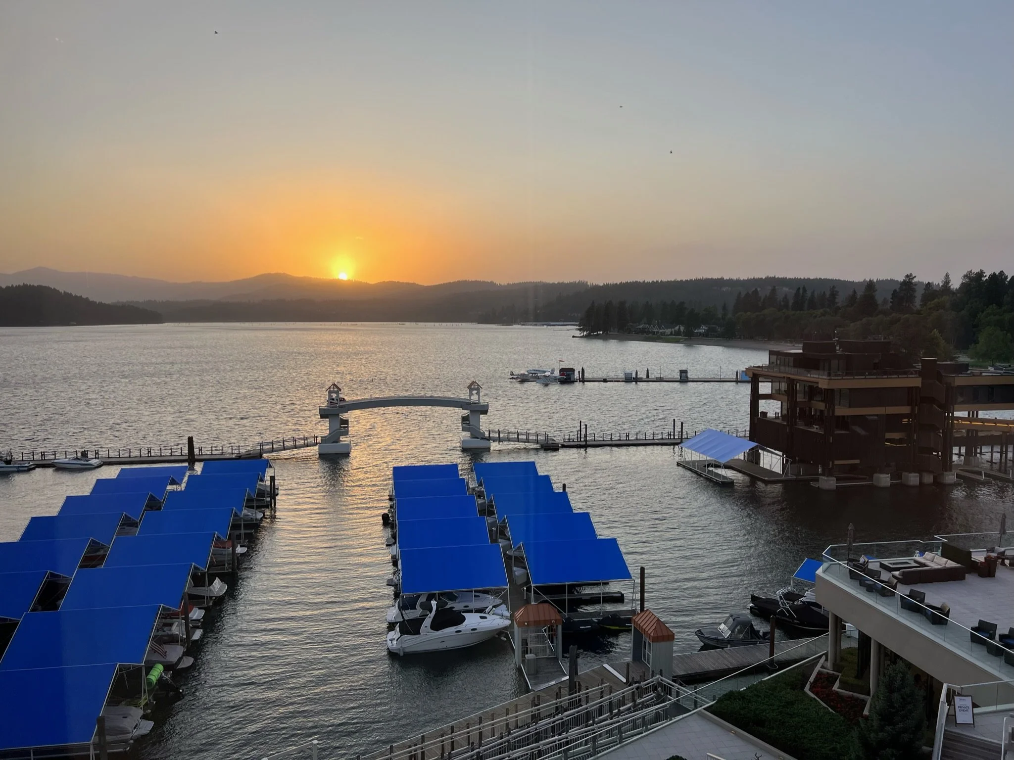 Sunset over a lake with boats docked at a marina, blue canopies covering some boats, a pier, and a building to the right, with mountains and trees in the background.