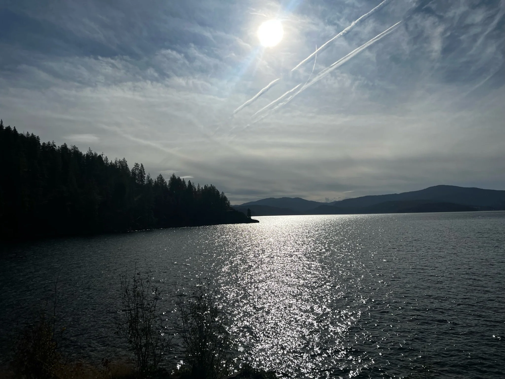 Sun shining over a lake with sparkling water, surrounded by forested hills and mountains in the distance, under a partly cloudy sky.