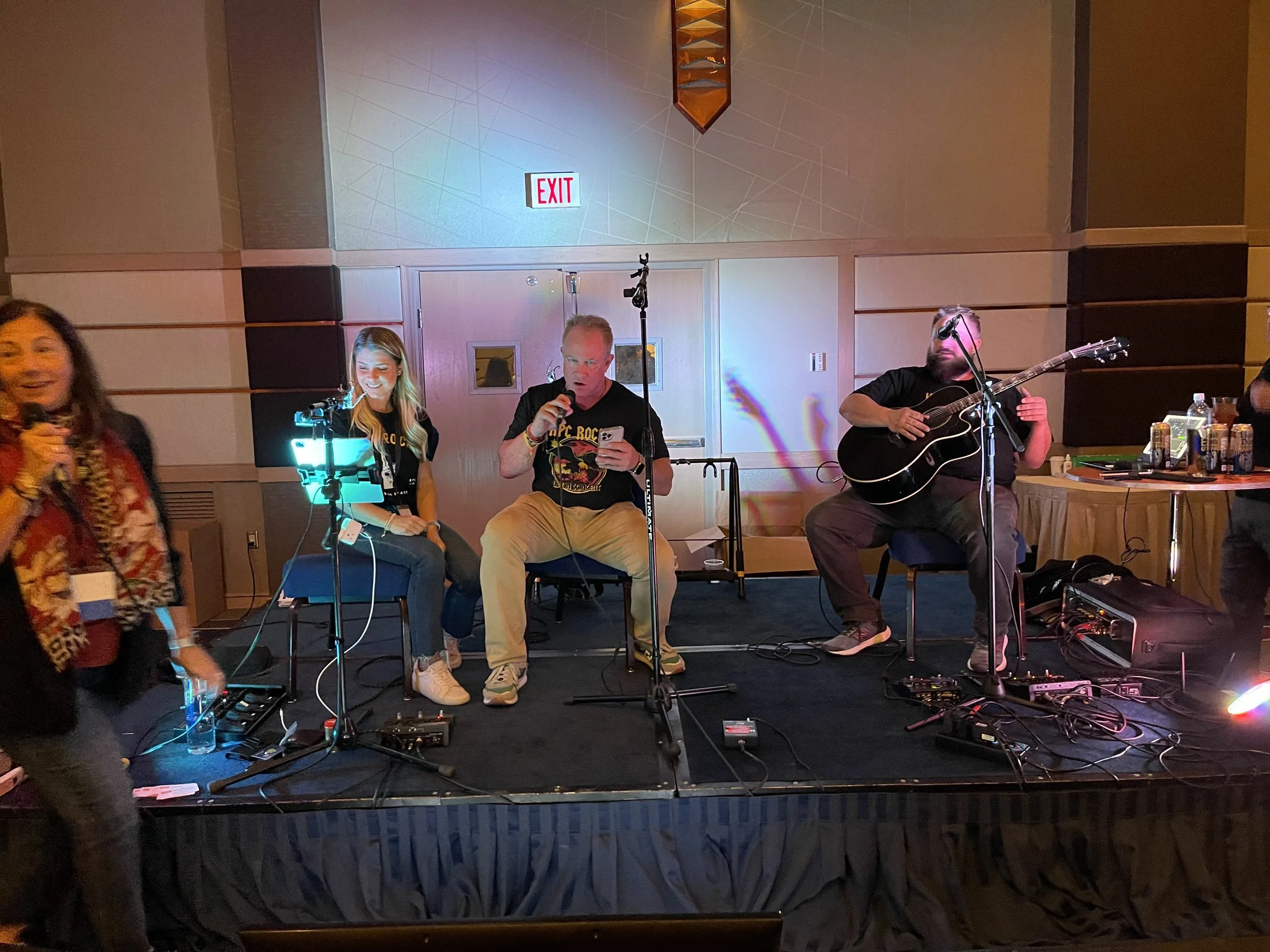 Four people in a room with a small stage, with three musicians sitting on chairs and a woman standing to the left holding a microphone. The musicians have instruments, including a guitar. On the right, there are drinks and equipment on a table.