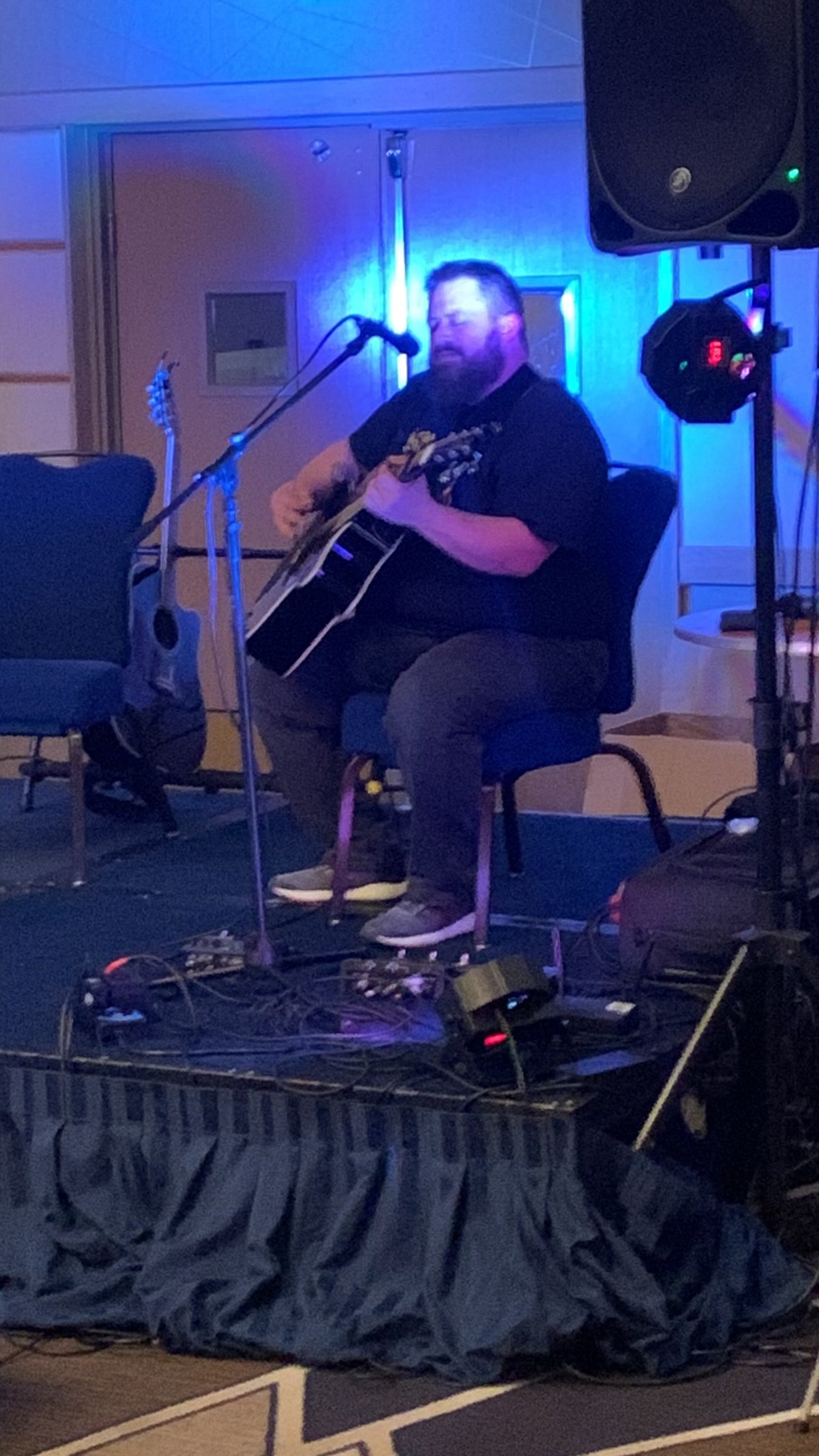 A man with a beard playing an acoustic guitar on stage with blue stage lighting.