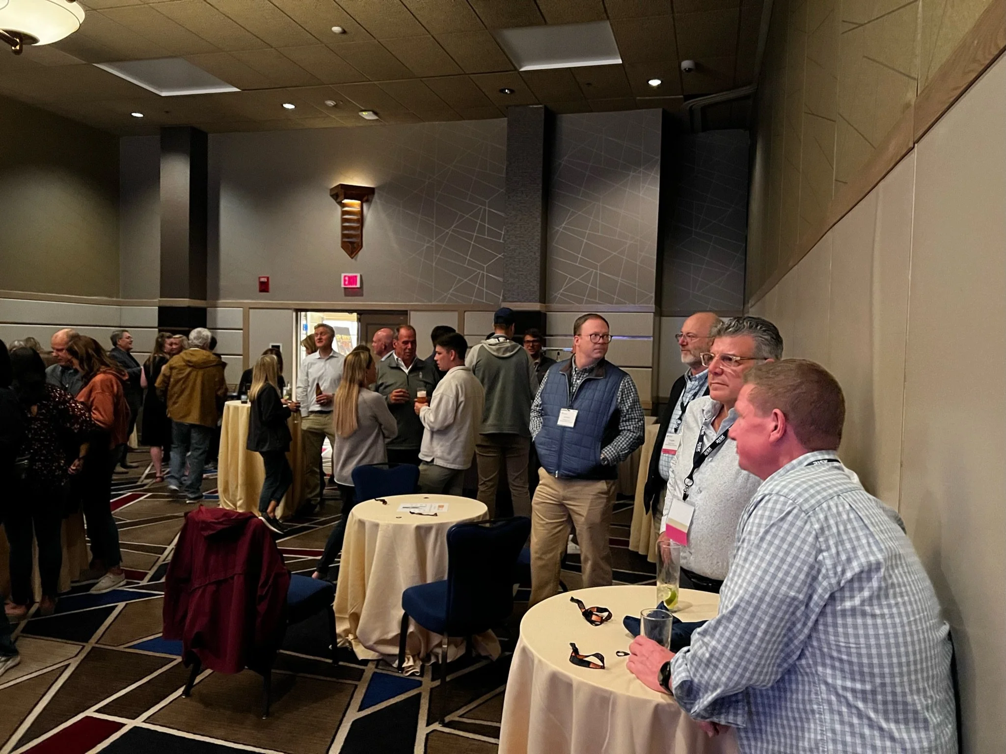 A group of people at a networking event in a conference room with round tables and beige walls.