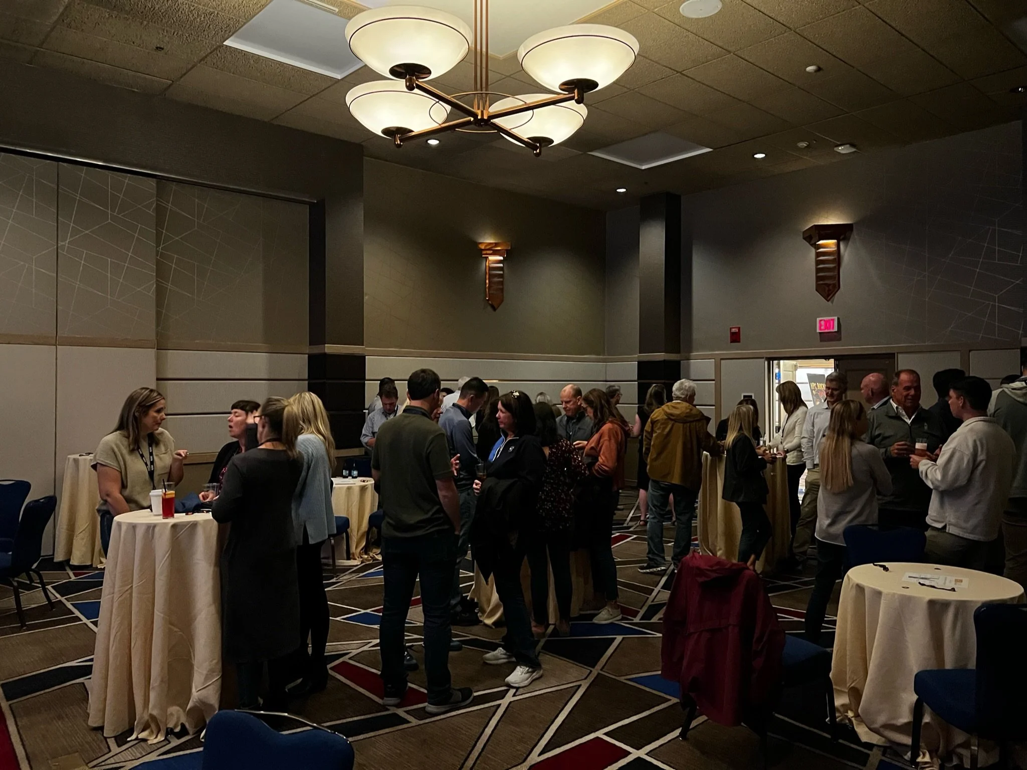 People socializing and networking at a conference or event in a large banquet hall with round tables and high ceilings.