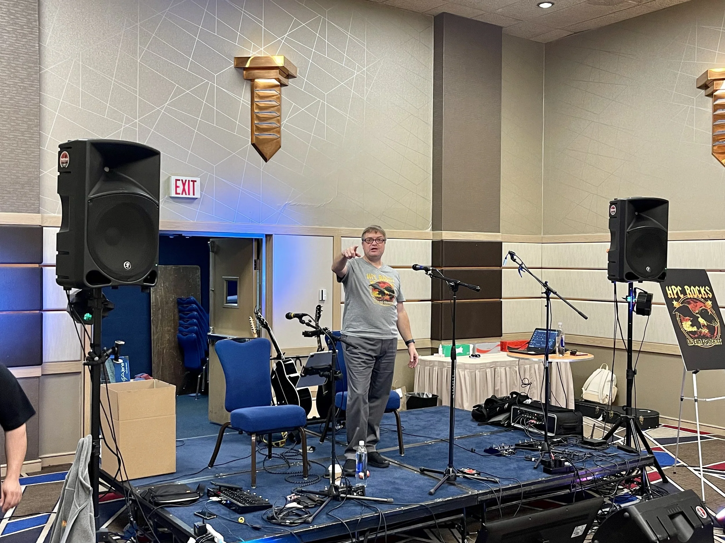 Man standing on a stage with microphone, surrounded by sound equipment, speakers, chairs, guitars, and a table with DJ equipment at an indoor event.