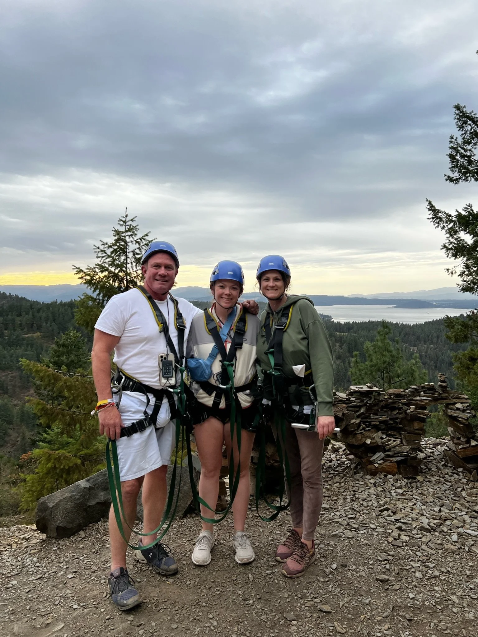 Three people wearing helmets and harnesses standing together outdoors with a scenic view of trees, water, and mountains in the background.
