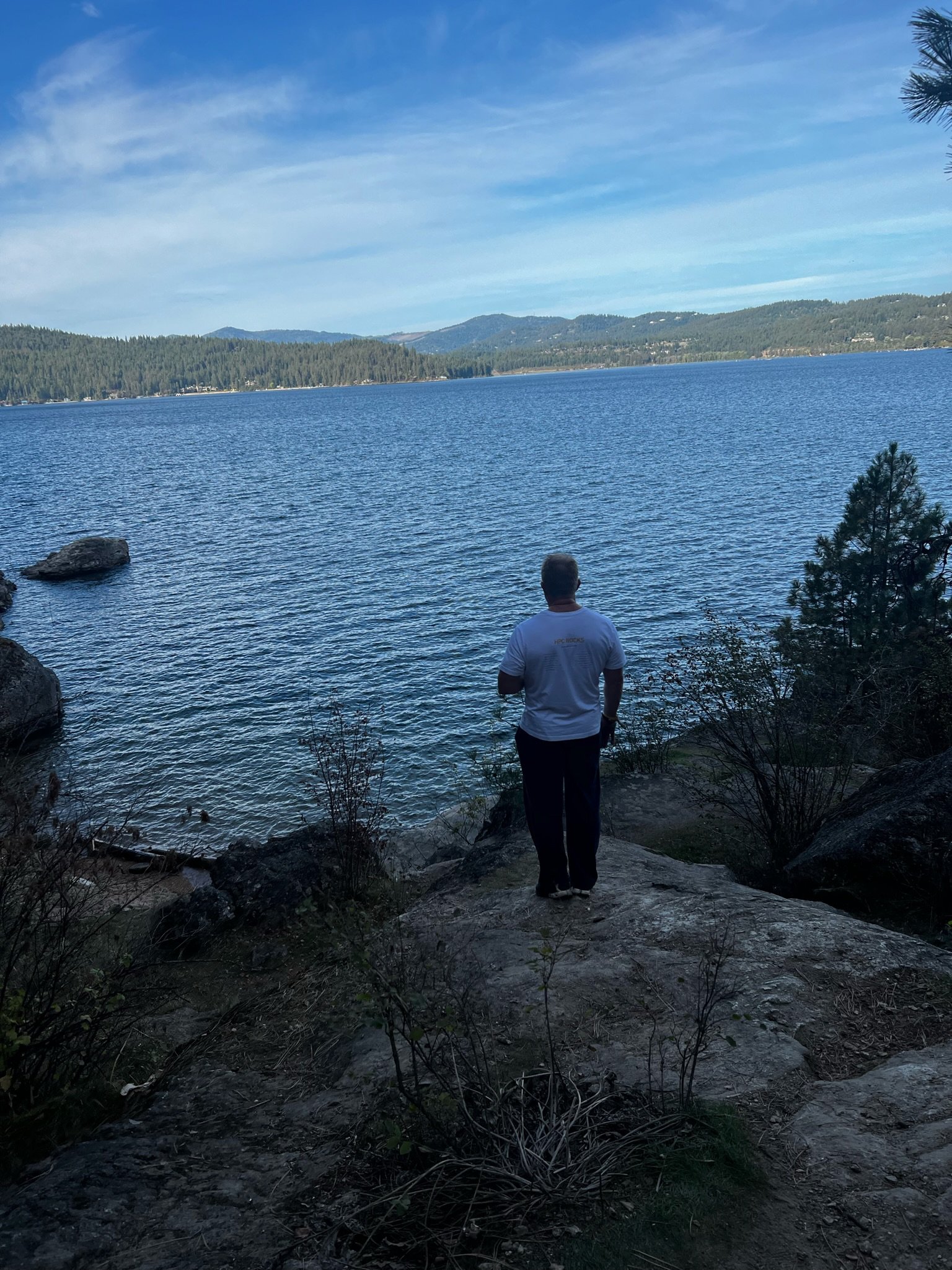 A person standing on a rocky shoreline, looking out over a large, calm lake surrounded by forested hills under a clear blue sky.