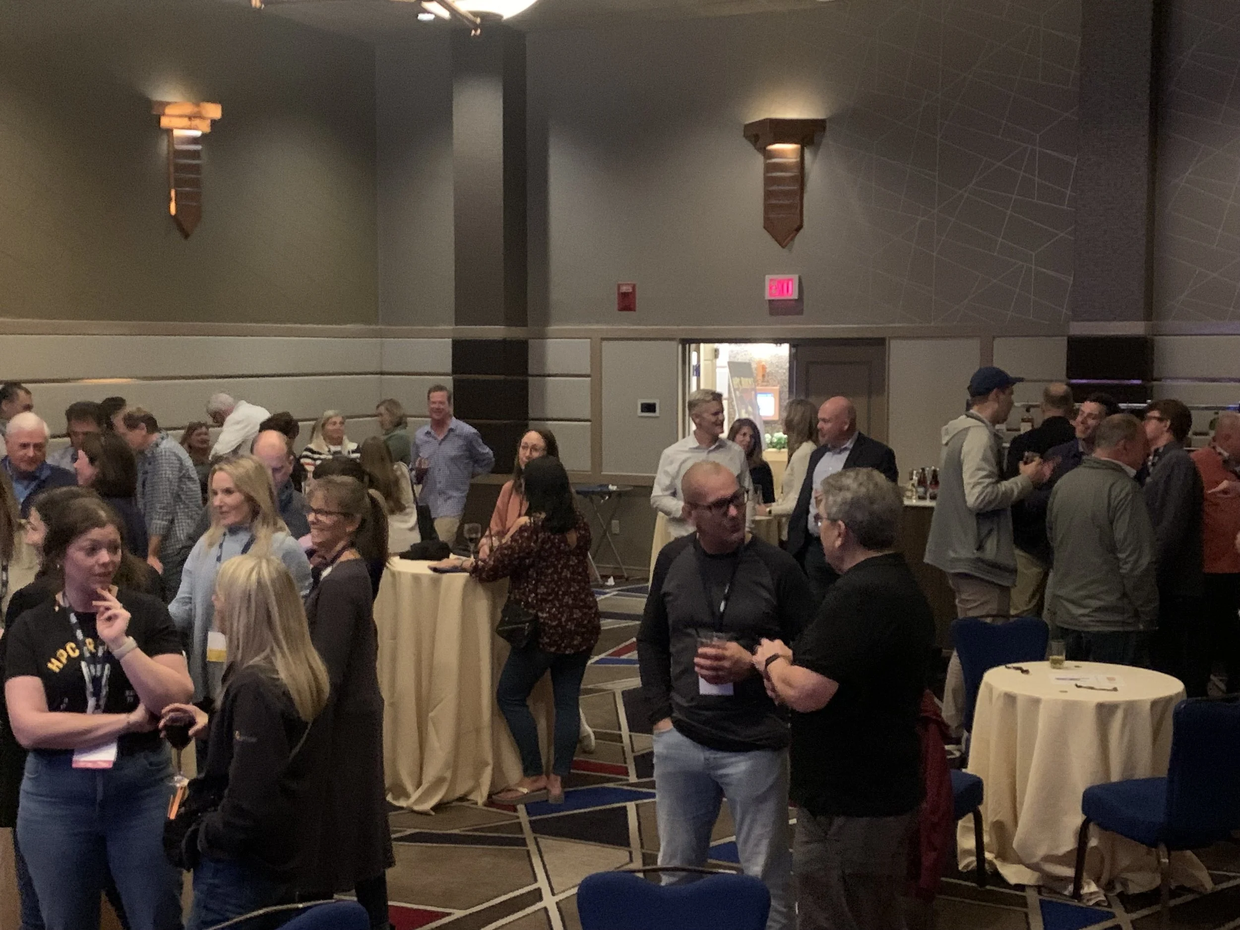 People socializing at a cocktail party in a conference hall with cocktail tables, some holding drinks, and engaged in conversation.