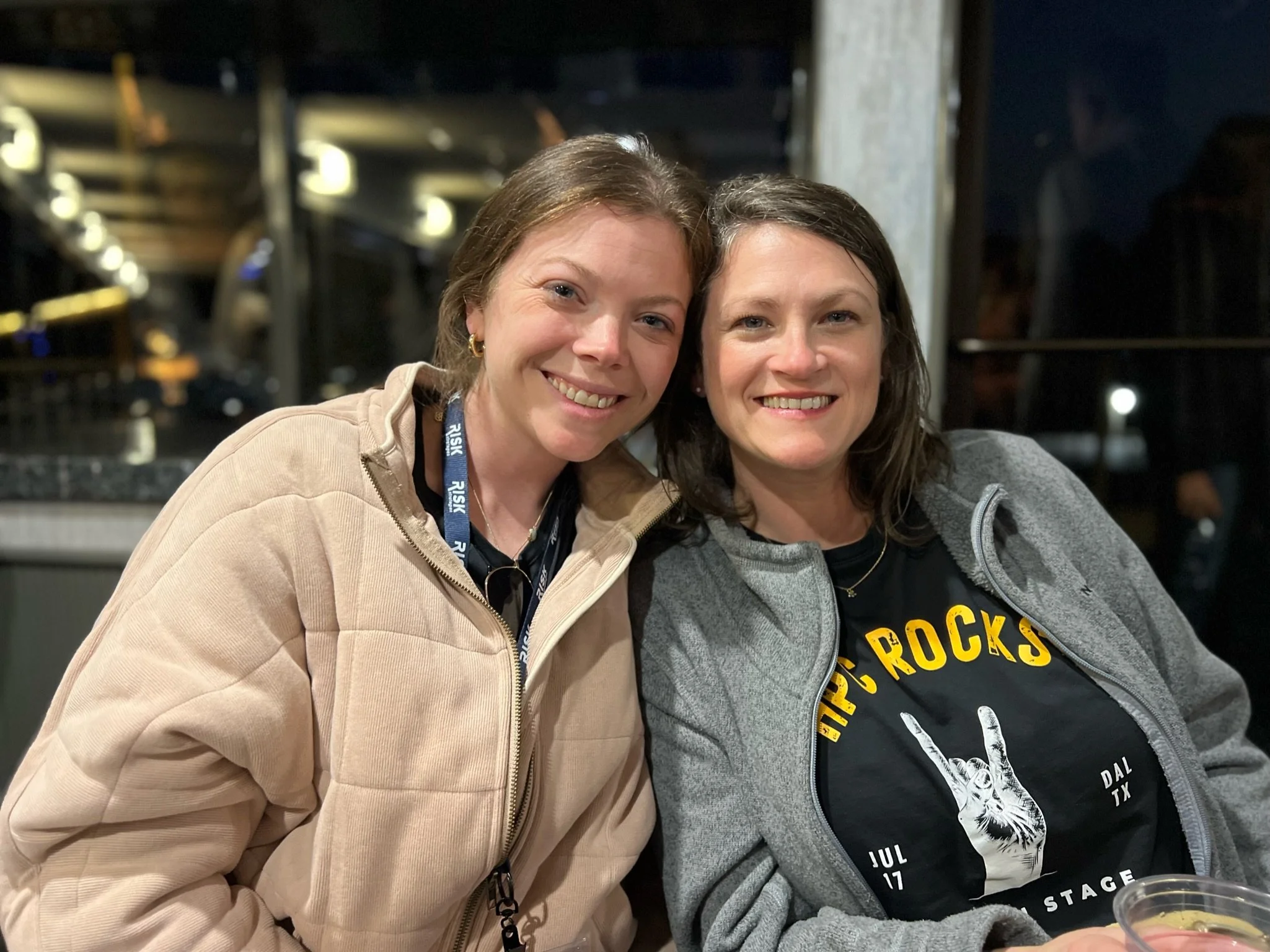 Two women sitting together indoors at night, smiling at the camera. One wearing a tan jacket with a black lanyard, the other wearing a gray hoodie over a black t-shirt with yellow and white lettering and a graphic of a hand making a rock and roll sig