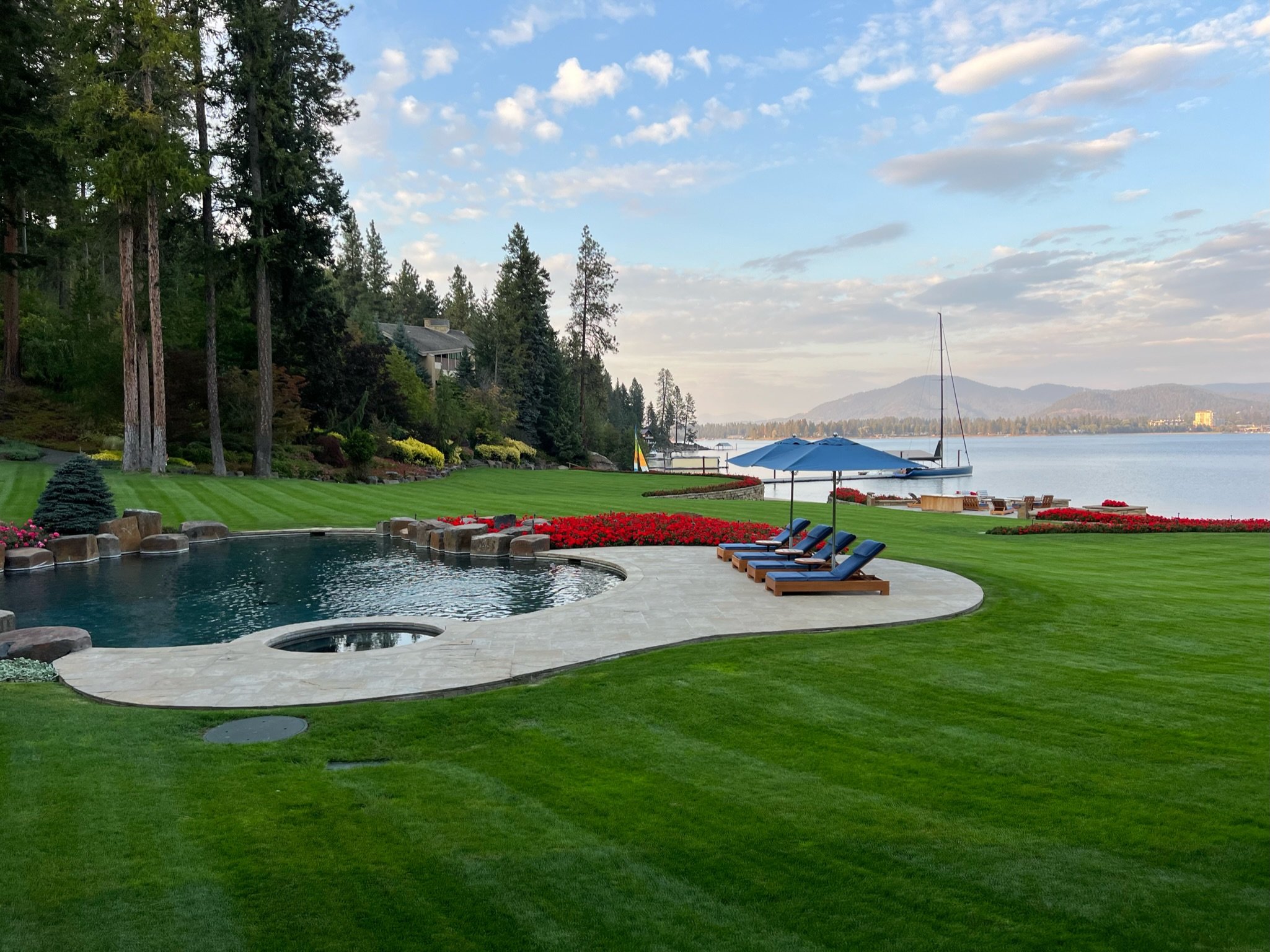 A luxurious backyard with a hot tub, green lawn, red flowers, and a view of the lake with a sailboat and mountains in the distance.