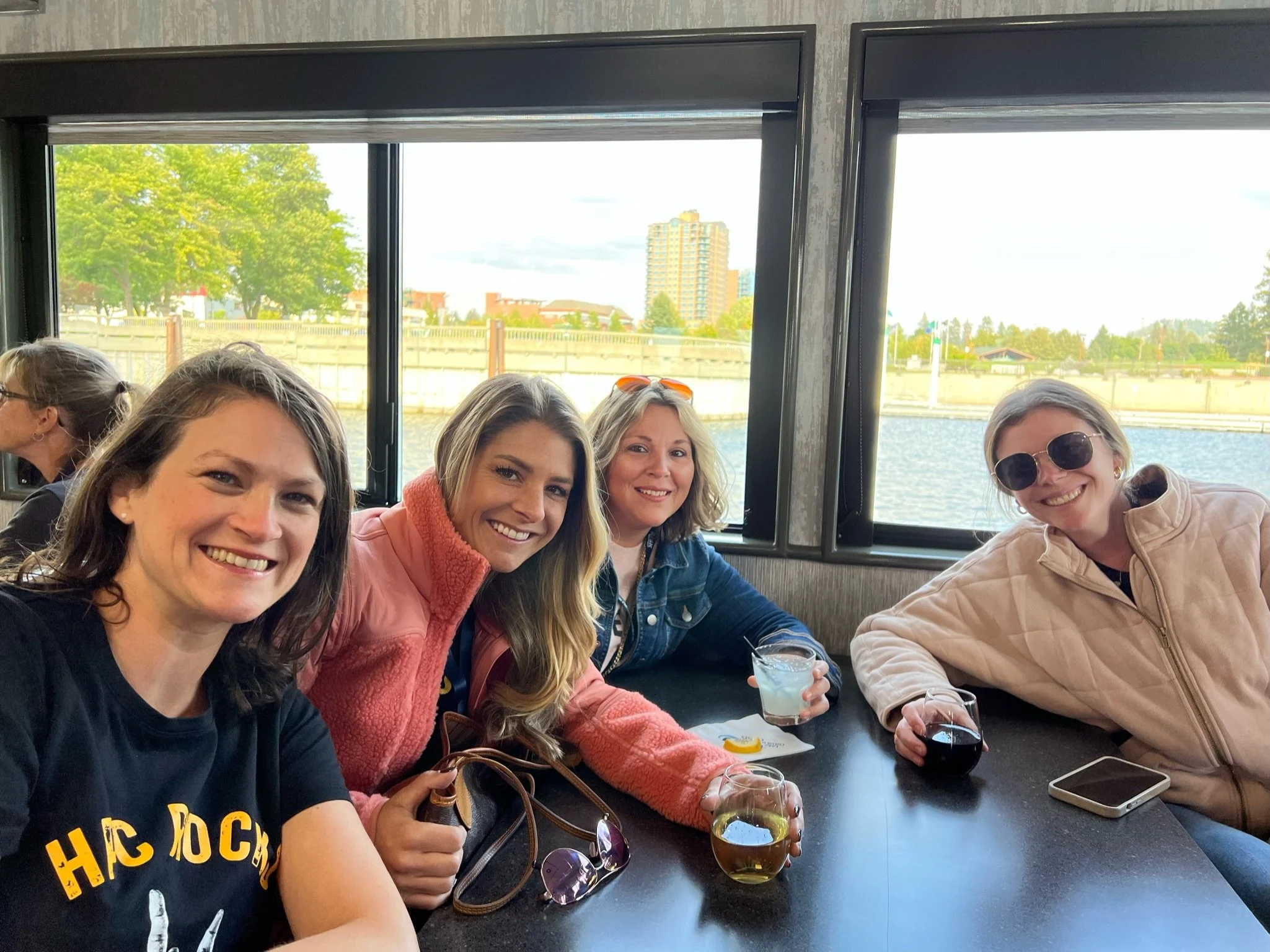 Four women sitting at a table near a window with water view, smiling and holding drinks, outdoor scene with trees and a city building in background.