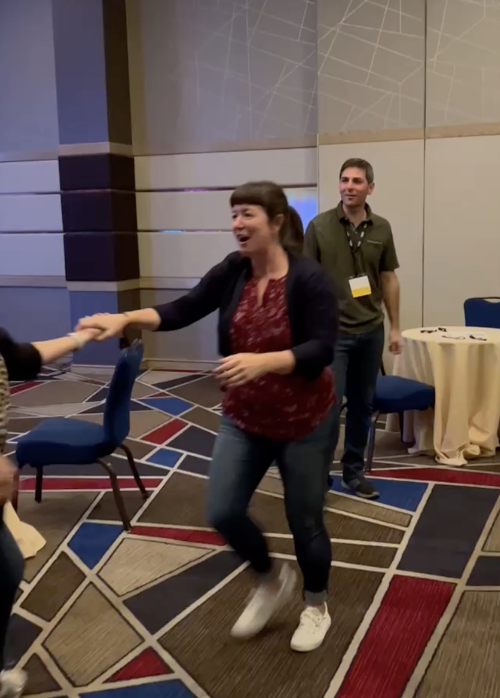 Two women and a man are in a conference room, with the women dancing and smiling while holding hands. The man stands nearby, smiling, and wearing a conference badge around his neck.