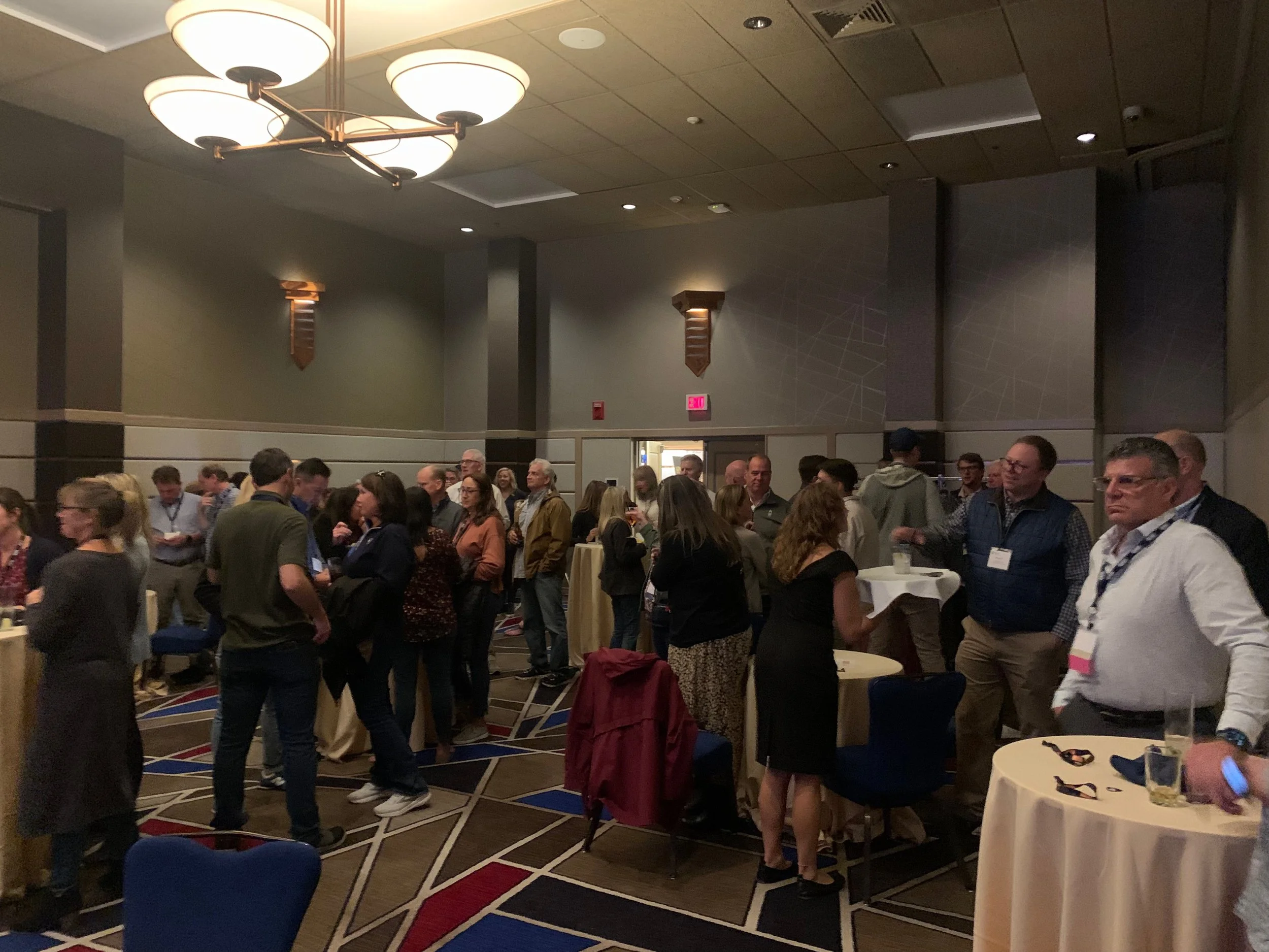 A group of people gathered in a conference room, standing around high-top tables, engaging in conversation at a professional networking event or conference.