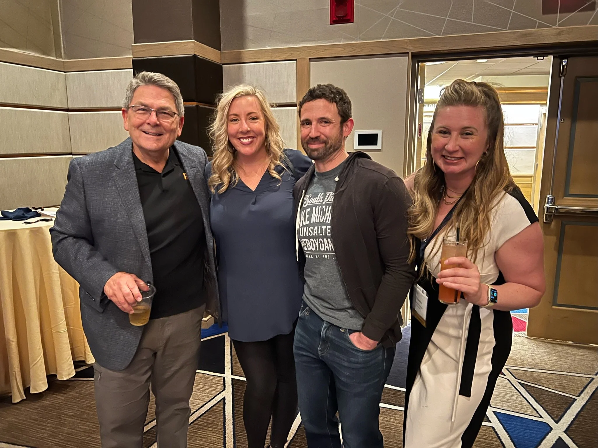 Four people standing together at an indoor event, smiling, with drinks in their hands.