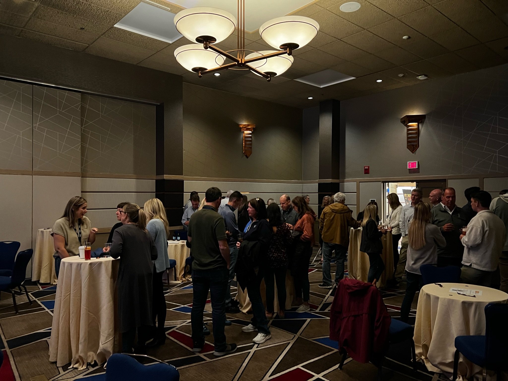 People mingling at a corporate networking event in a conference room with high ceilings, modern lighting, and a geometric rug pattern.