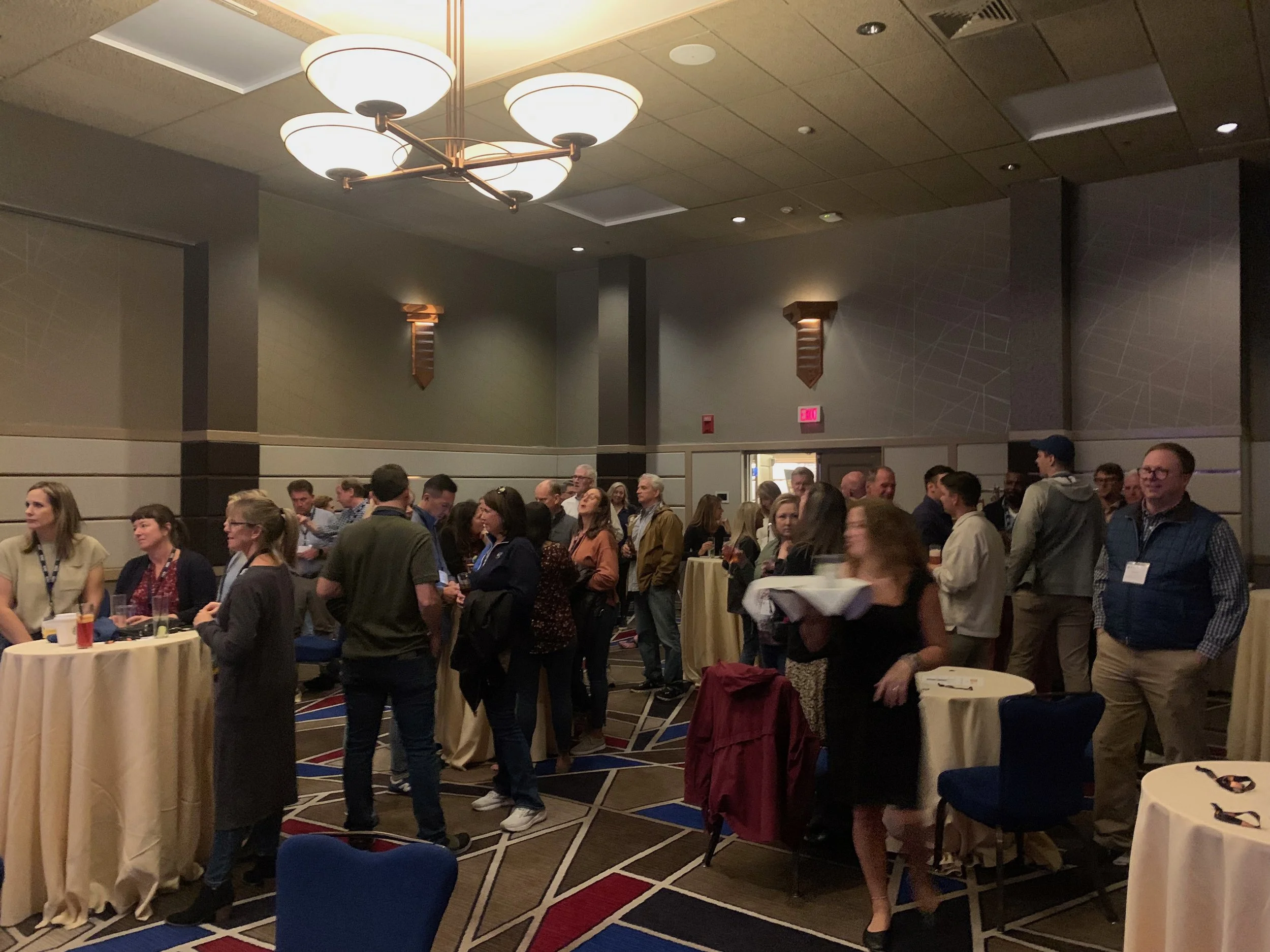 People gathered in a conference room with standing tables, some holding drinks or food plates, engaging in conversations under warm lighting and modern decor.
