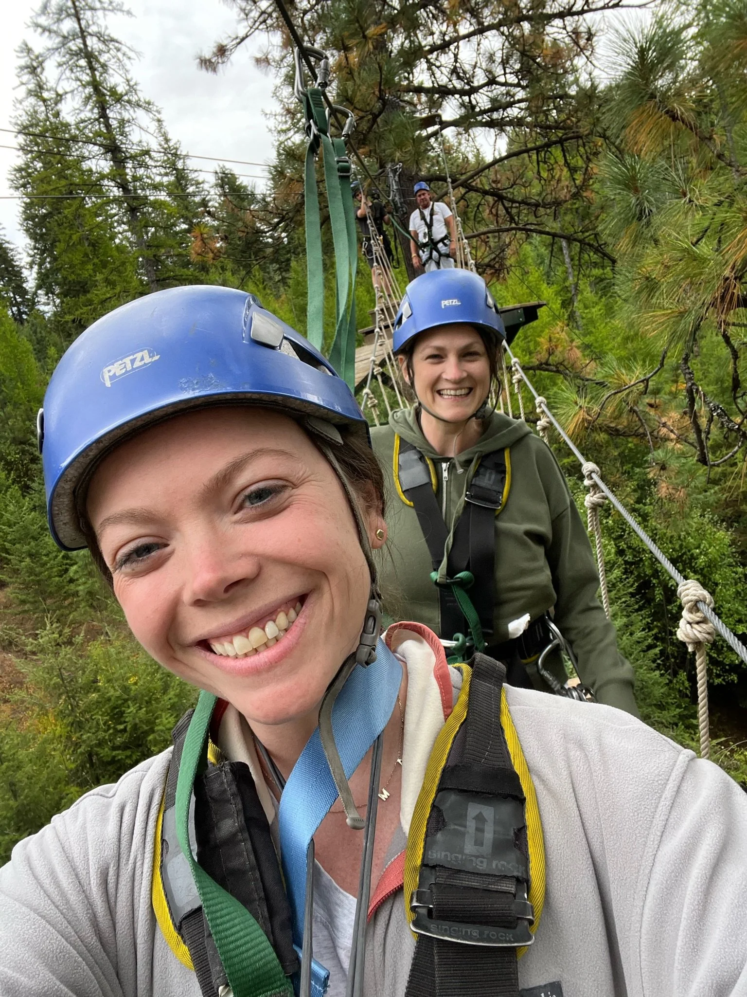 Three people, two women and one man, wearing helmets and harnesses, are on a suspension bridge in a forested area with trees and cloudy sky in the background. They are smiling and appear to be enjoying an outdoor adventure or zip-lining activity.