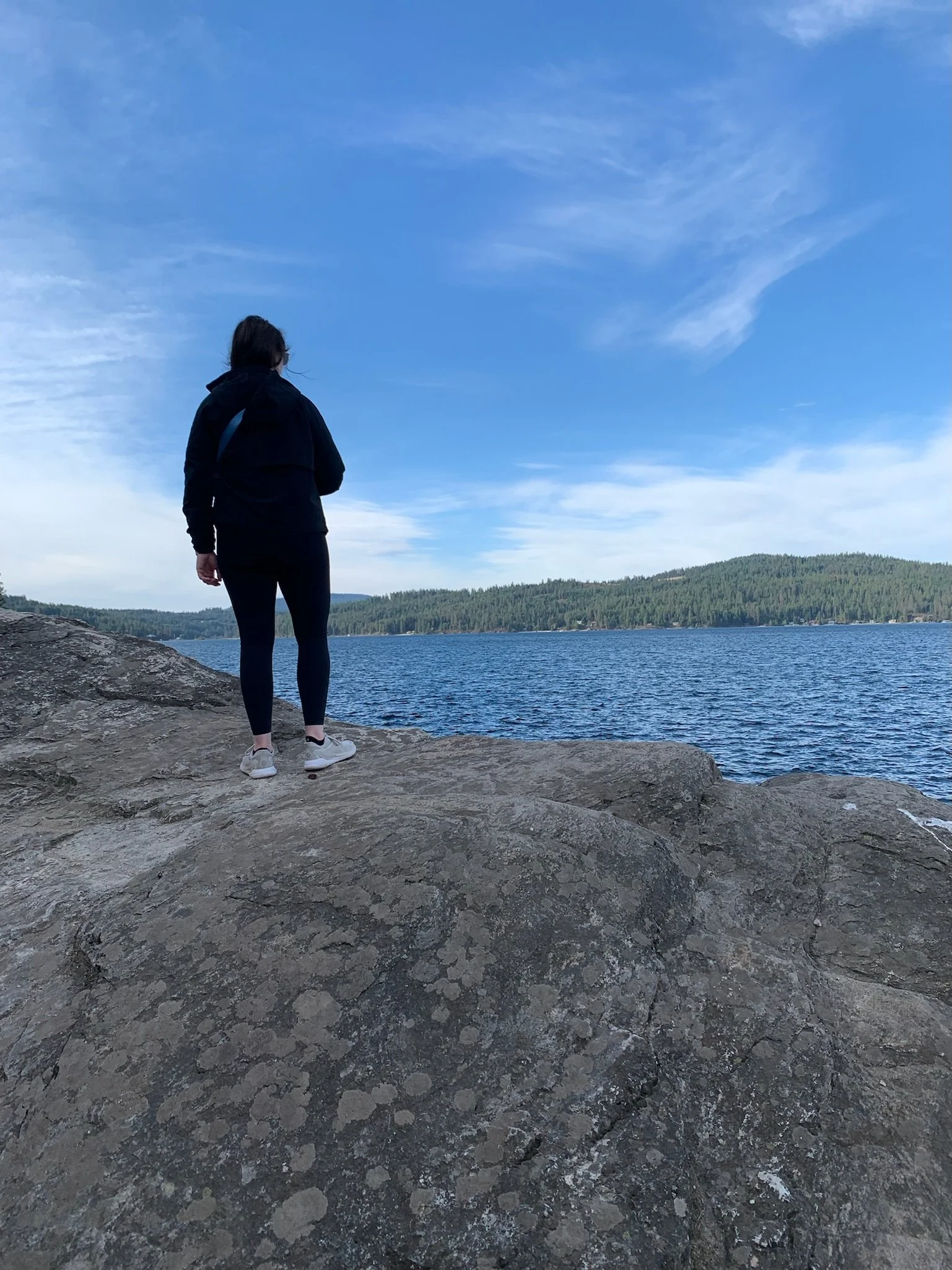 Person standing on large rocks by a lake, overlooking water with trees and hills in the distance under a partly cloudy sky.