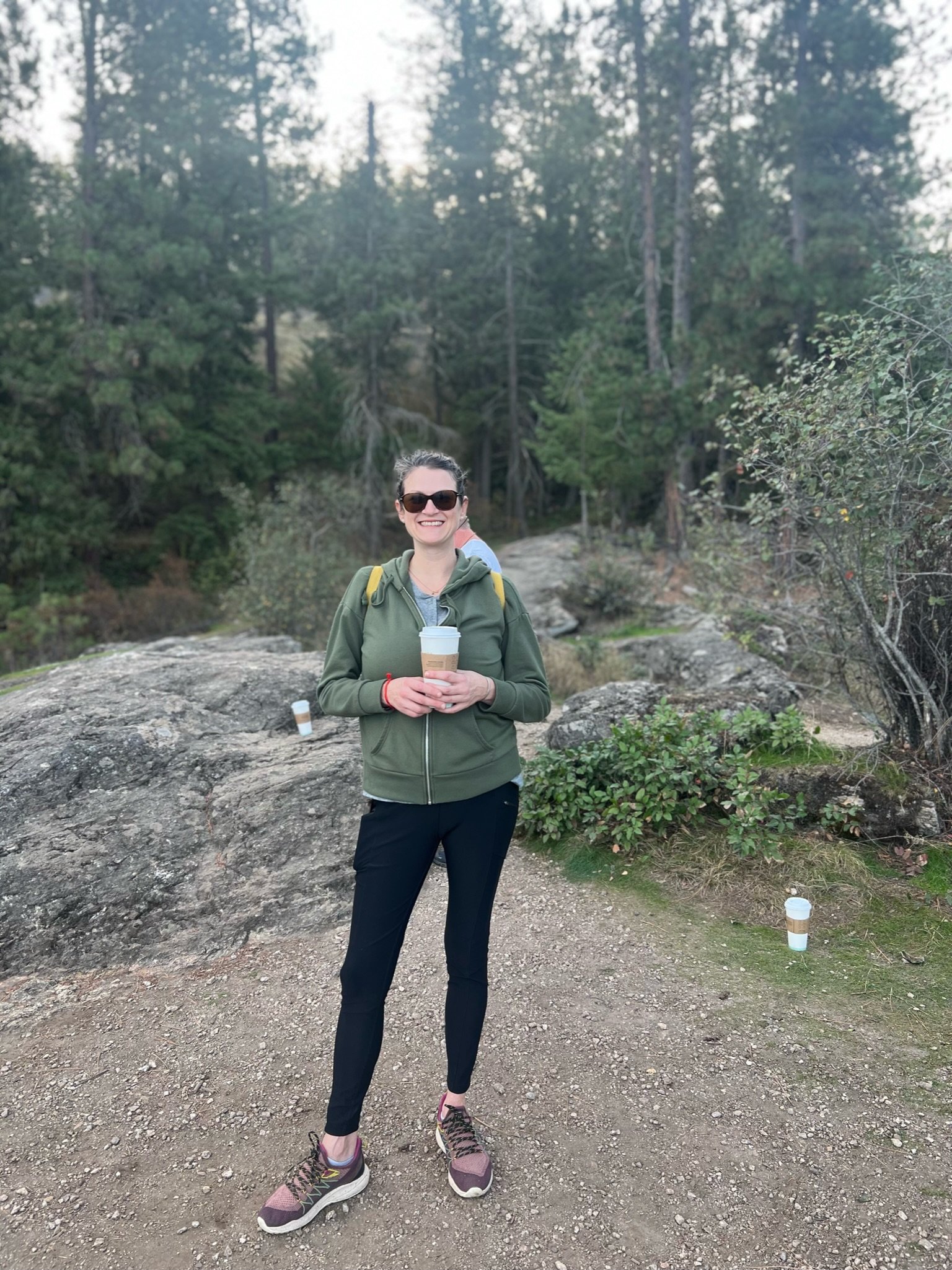 A woman in outdoor hiking attire, holding a coffee cup, standing on a dirt trail in a forested area with trees and rocks.