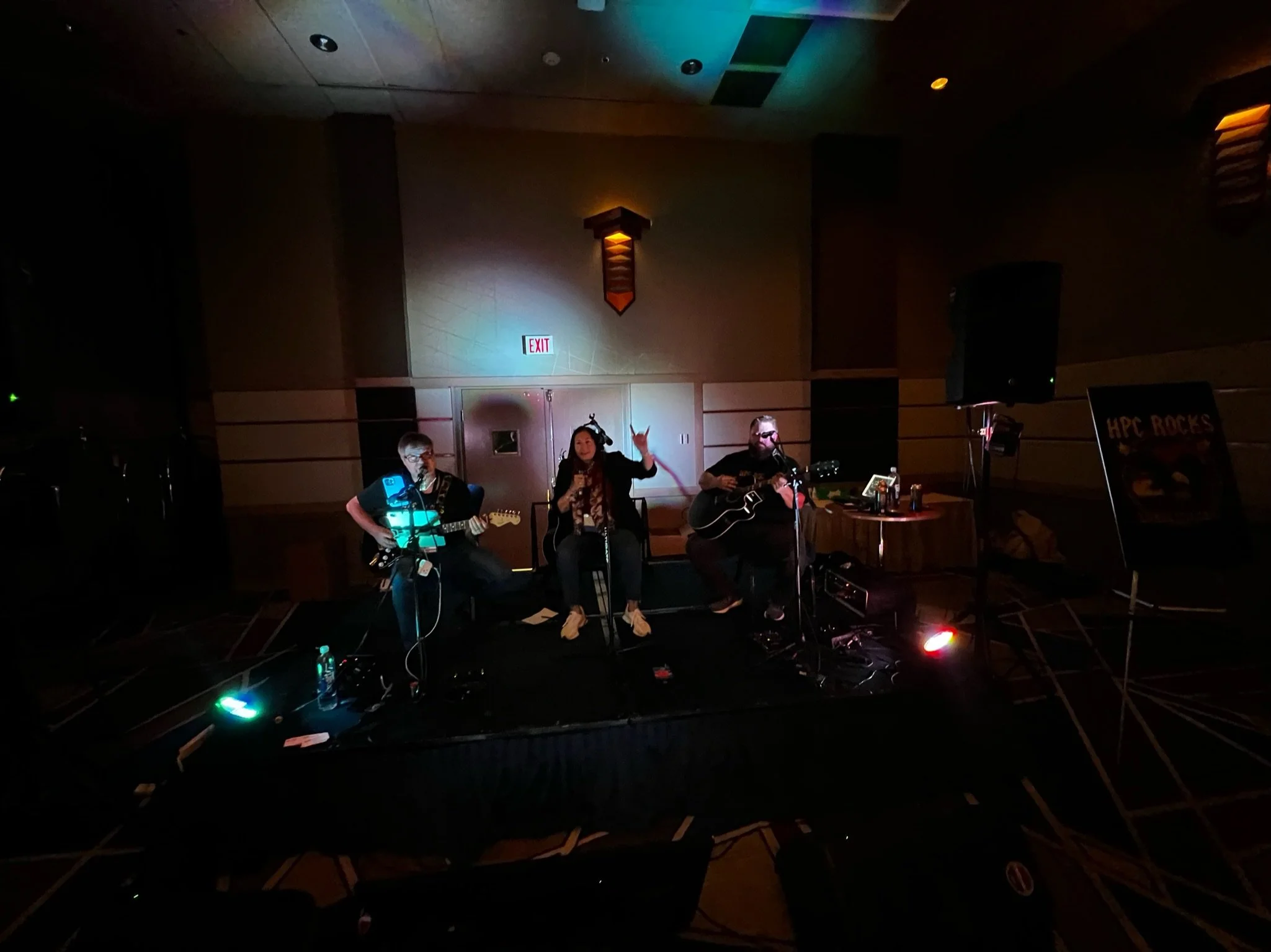 A live musical performance on a small stage with three musicians: two guitarists and a singer, in a dimly lit room with colorful stage lights, a sign that says 'HPC ROCKS' on the right, and an exit door in the background.