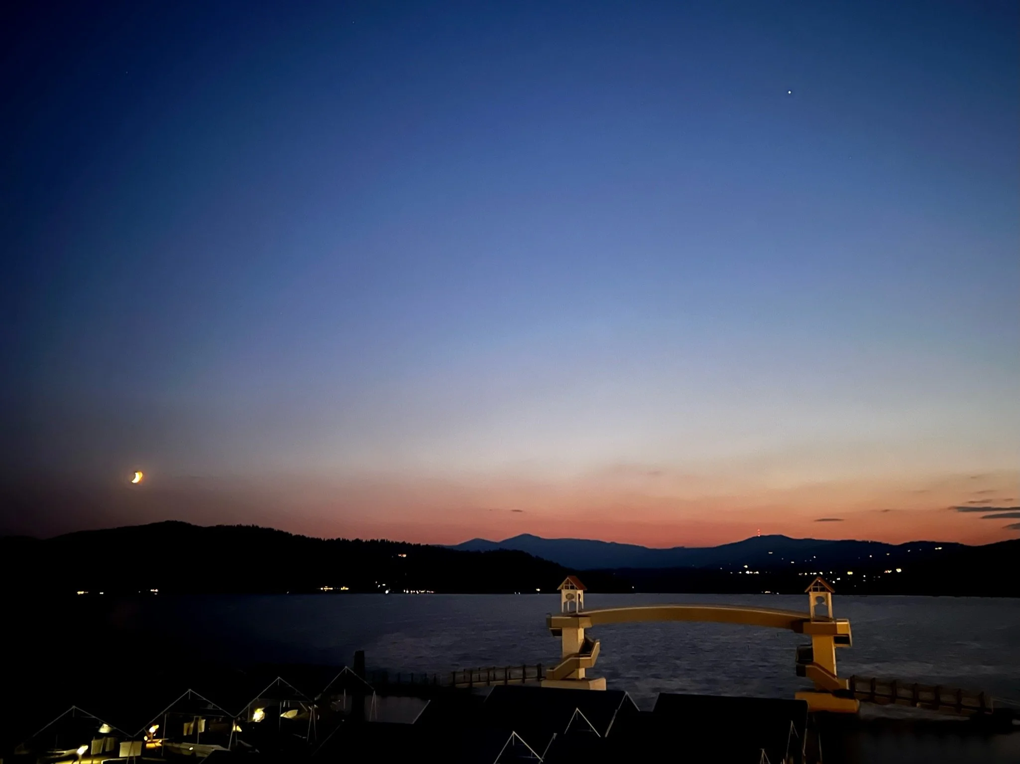 Dusk over a landscape with mountains, a crescent moon, and a bright planet or star in the sky. A footbridge over the water is visible in the foreground.