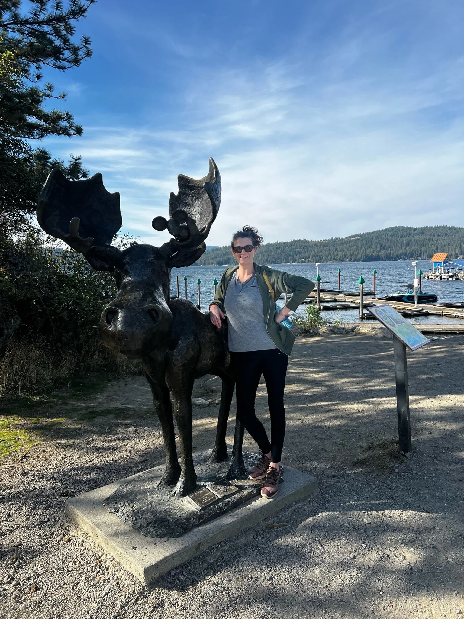 A woman standing next to a large moose sculpture with a cartoonish moose head on its antlers at a lakeside. The background features a lake with docks, boats, and wooded hills, under a partly cloudy sky.