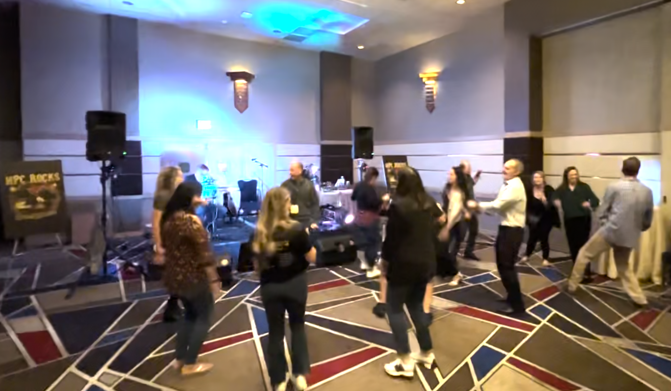 People dancing and socializing at an indoor event with stage and DJ setup.