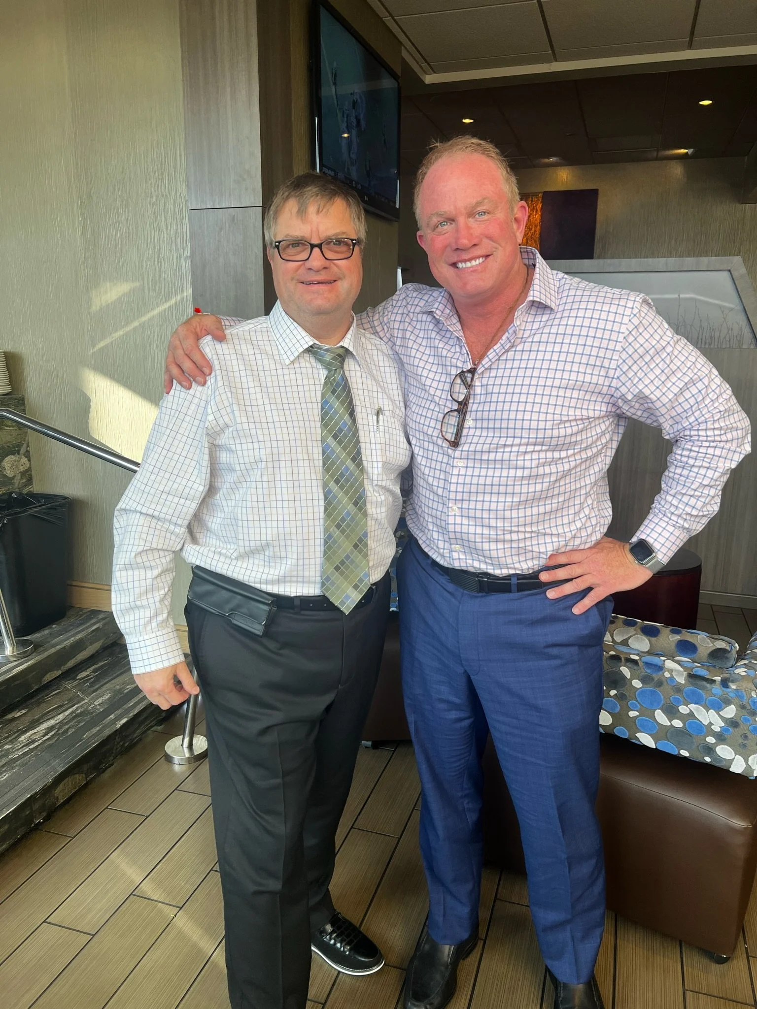 Two men standing indoors, smiling, with one man wearing glasses, a dress shirt, and a tie, and the other man in a checked shirt with sunglasses hanging on his collar, and a smartwatch on his wrist.