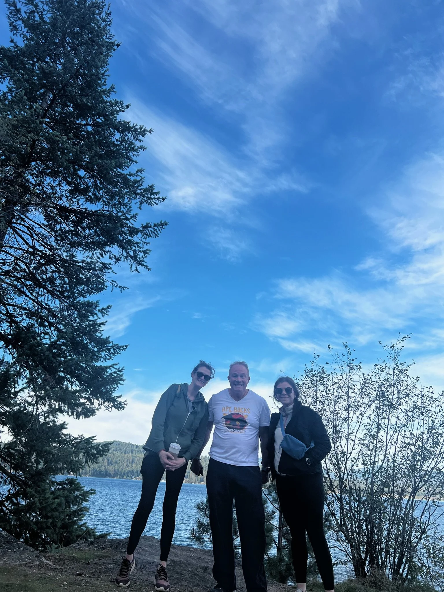 Three people smiling and standing outdoors near a lake with trees and a blue sky in the background.
