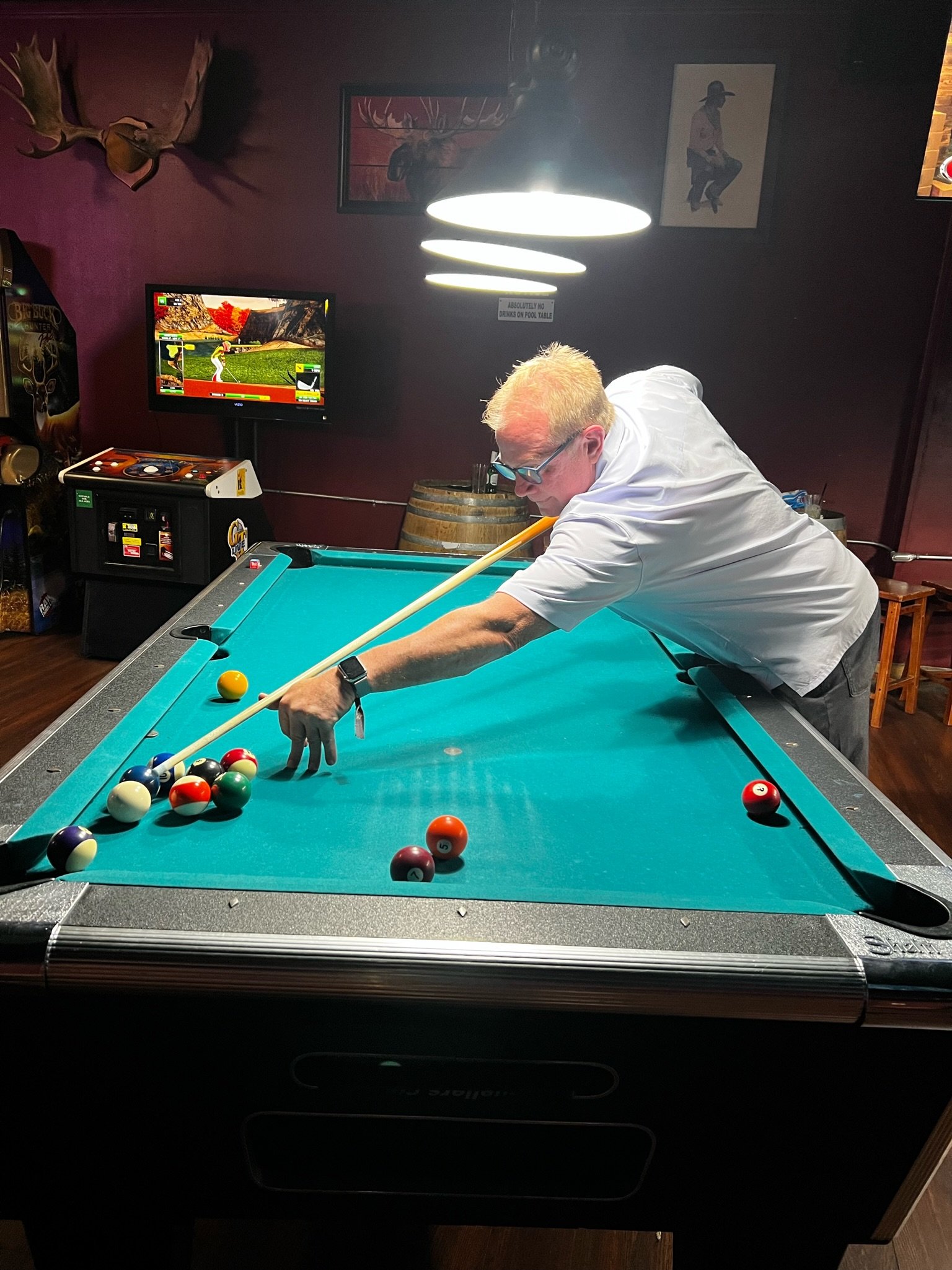 A man playing pool on a teal-colored billiards table in a dimly lit room. He is aiming at the balls with a cue stick. In the background, there is a television, a dartboard, and some wall art.