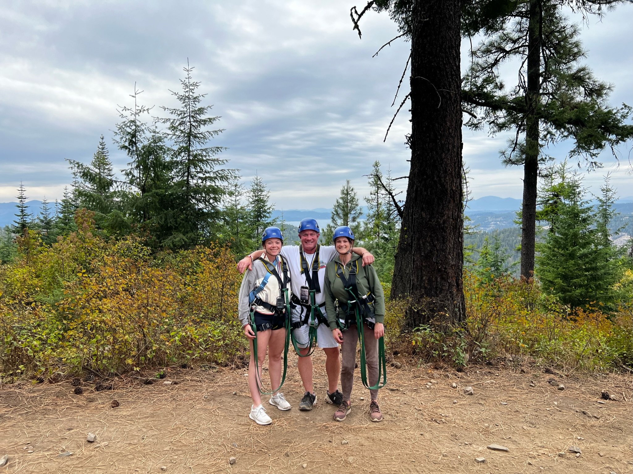 Three people in hiking gear and helmets standing together outdoors in a forest, smiling with arms around each other.