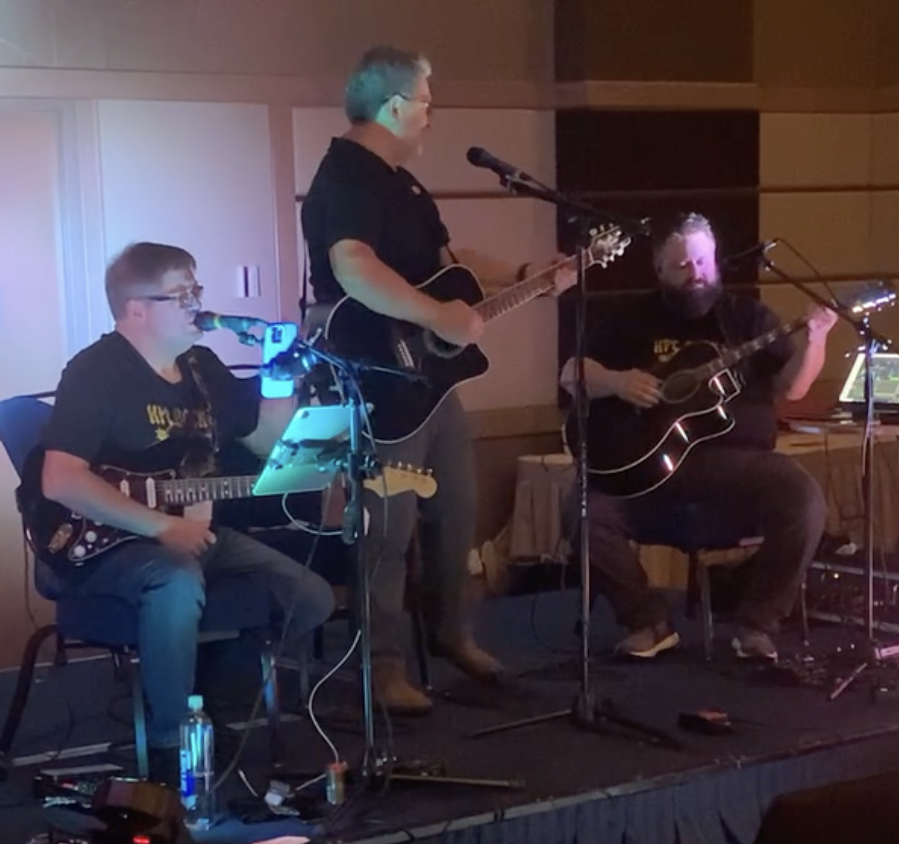 Three men playing guitars on a stage, one singing into a microphone, in a dimly lit room with wood-paneled walls.