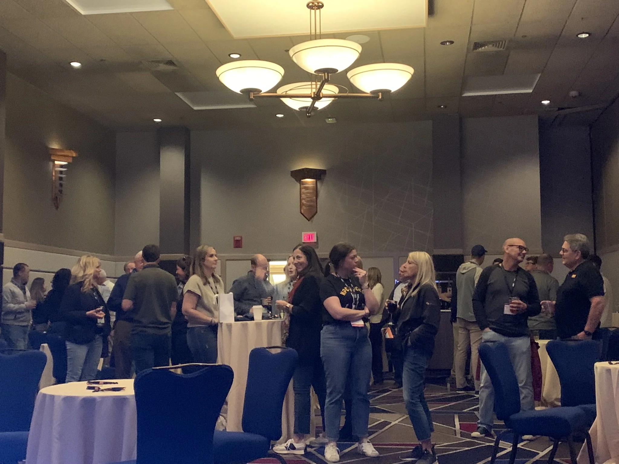 People mingling at a social event in a large, dimly lit banquet hall with round tables and blue chairs. Some individuals are holding drinks and engaging in conversation.
